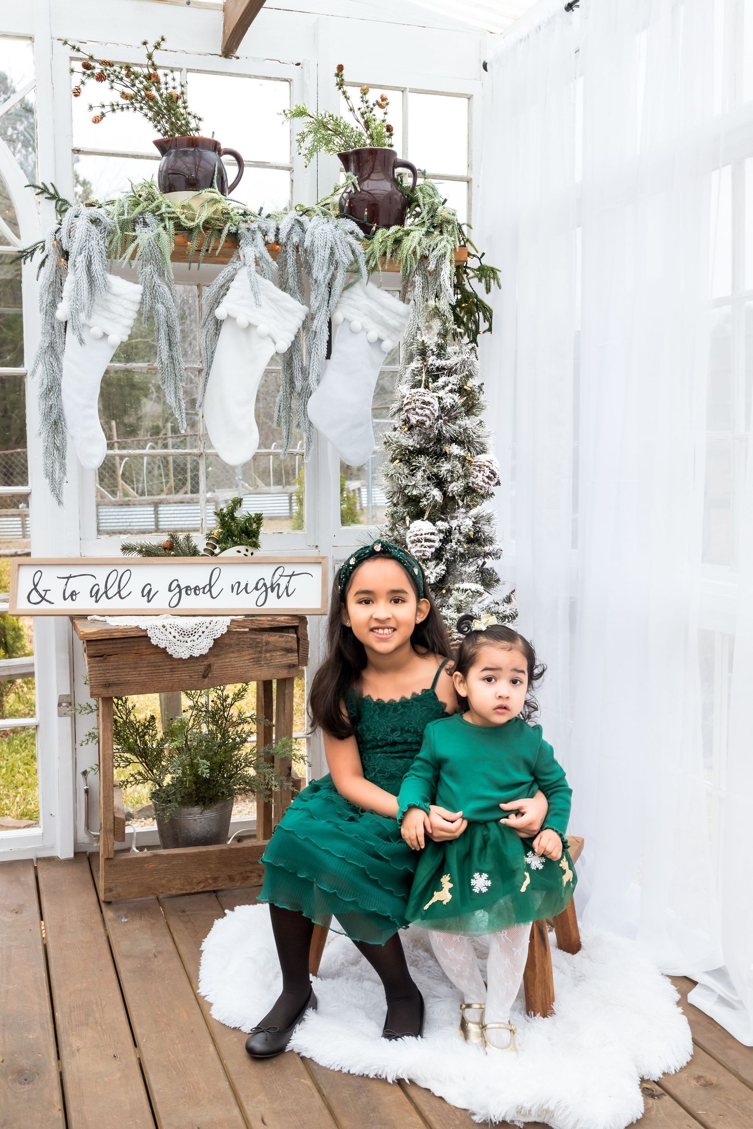 Two children in matching emerald green sweaters sit together on a white bench with holiday decorations.
