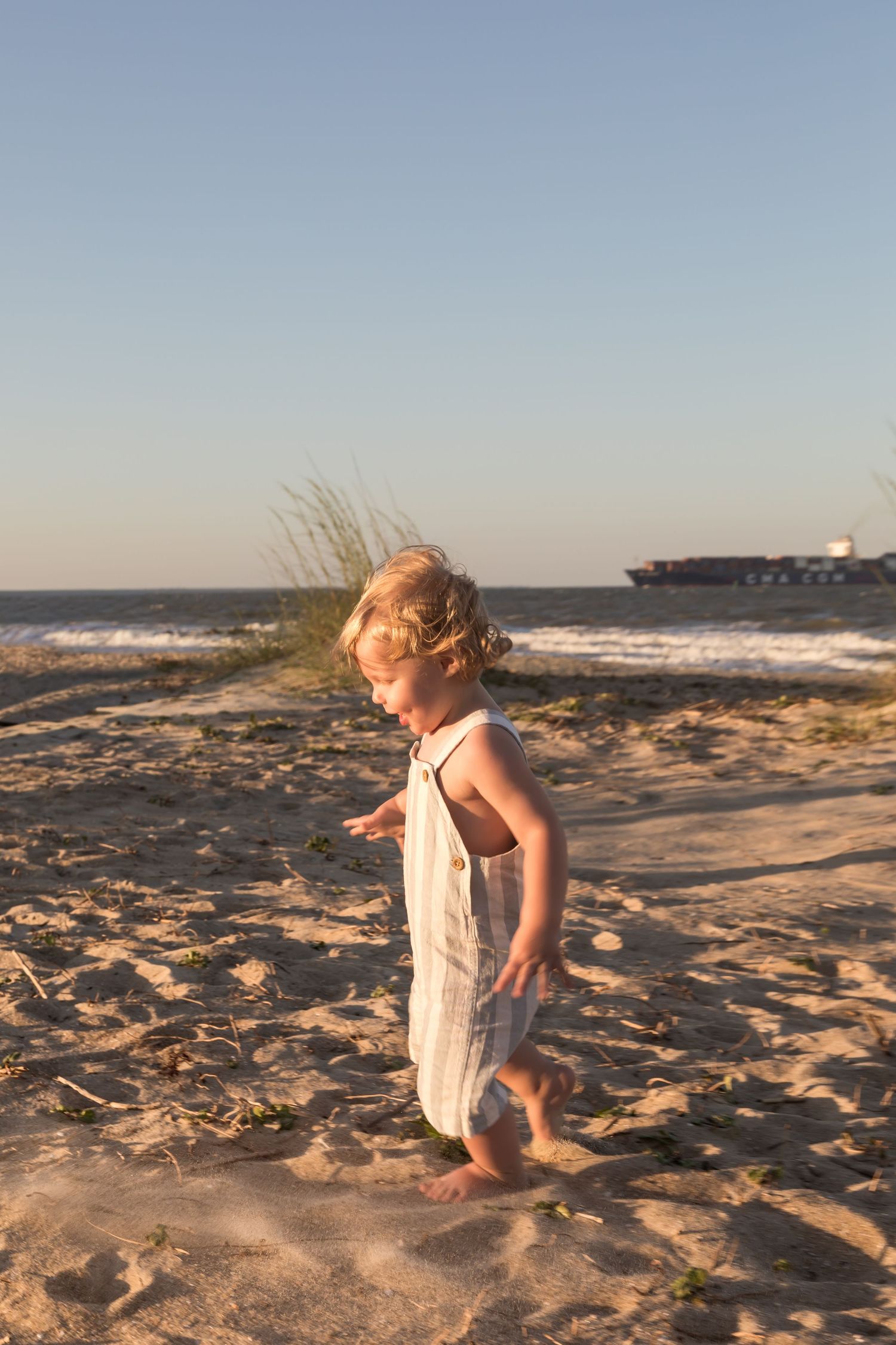 Young blonde toddler girl in white sundress walking barefoot on sandy beach at golden hour sunset.