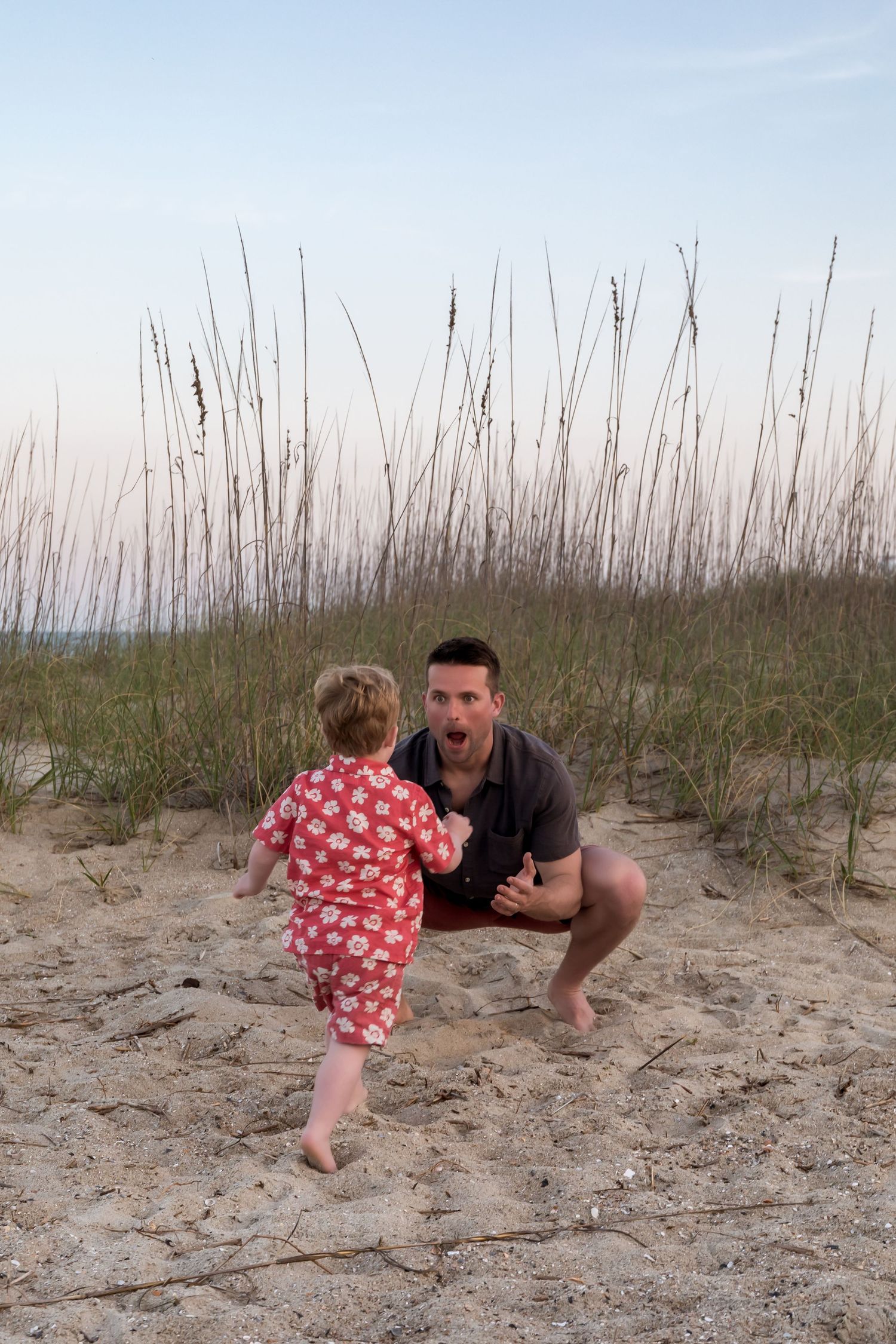 A father crouches on sandy beach dunes playfully greeting his toddler in red floral outfit near sea oats at dusk.