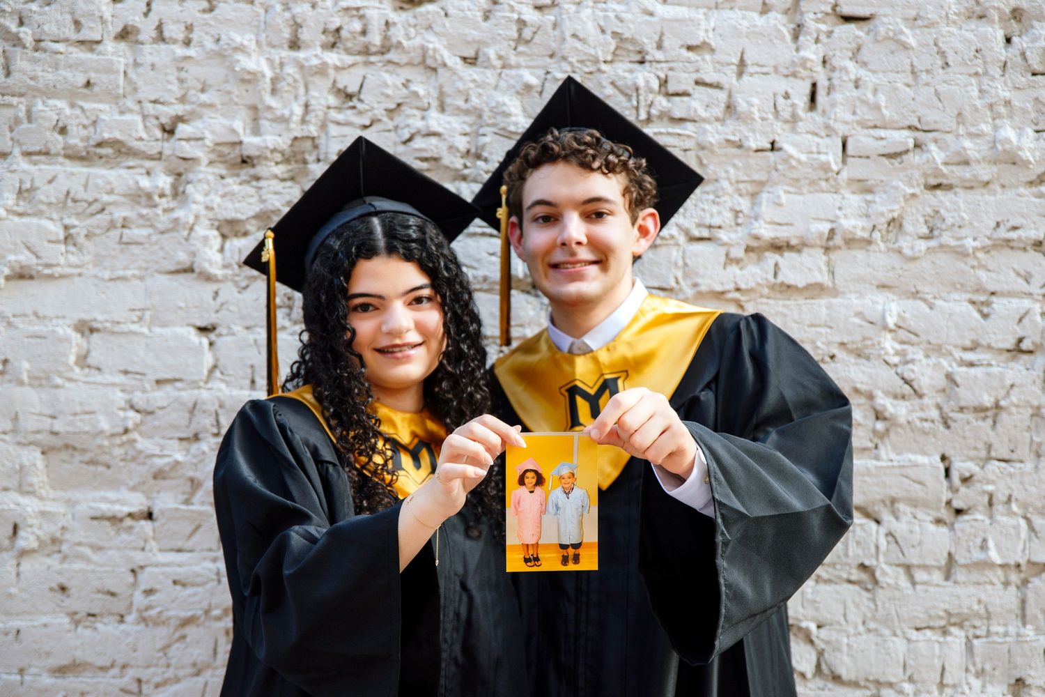 Graduates display their diplomas while celebrating in front of a textured brick wall.
