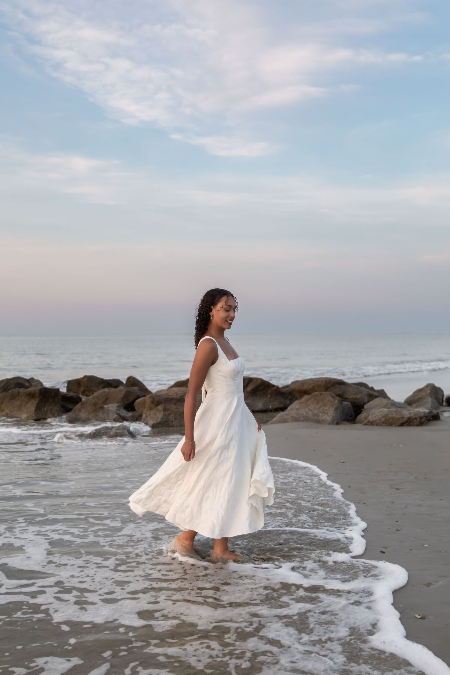 Woman in white dress walking barefoot on beach shore with rocks and ocean waves at sunset.