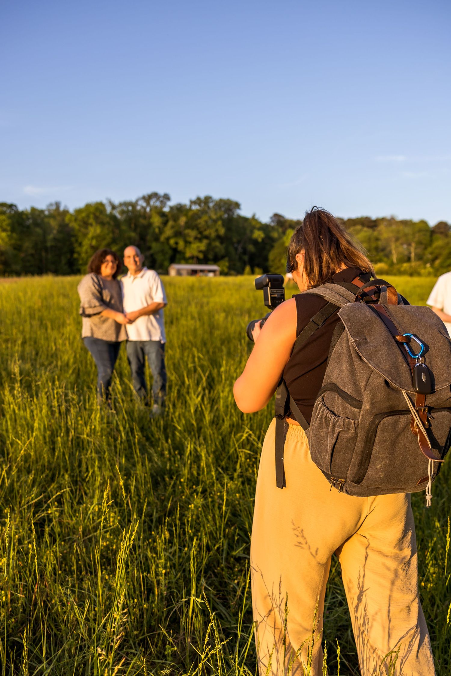 Photographer with backpack capturing couple in golden field during sunset.