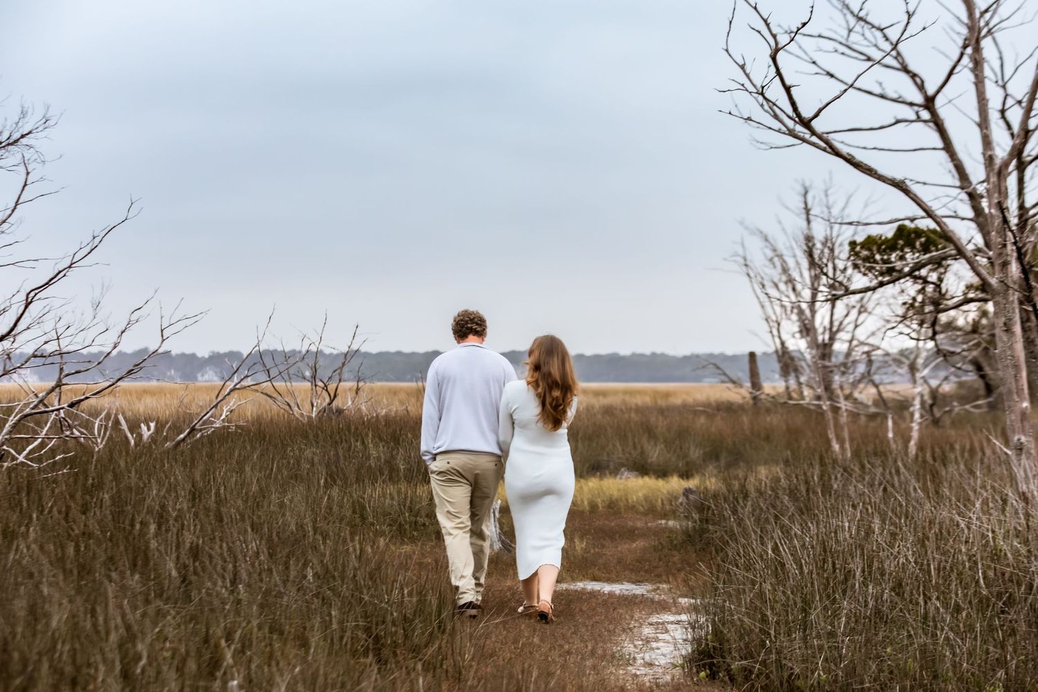 A couple walks hand in hand through tall beach grass along a scenic coastal path at sunset.