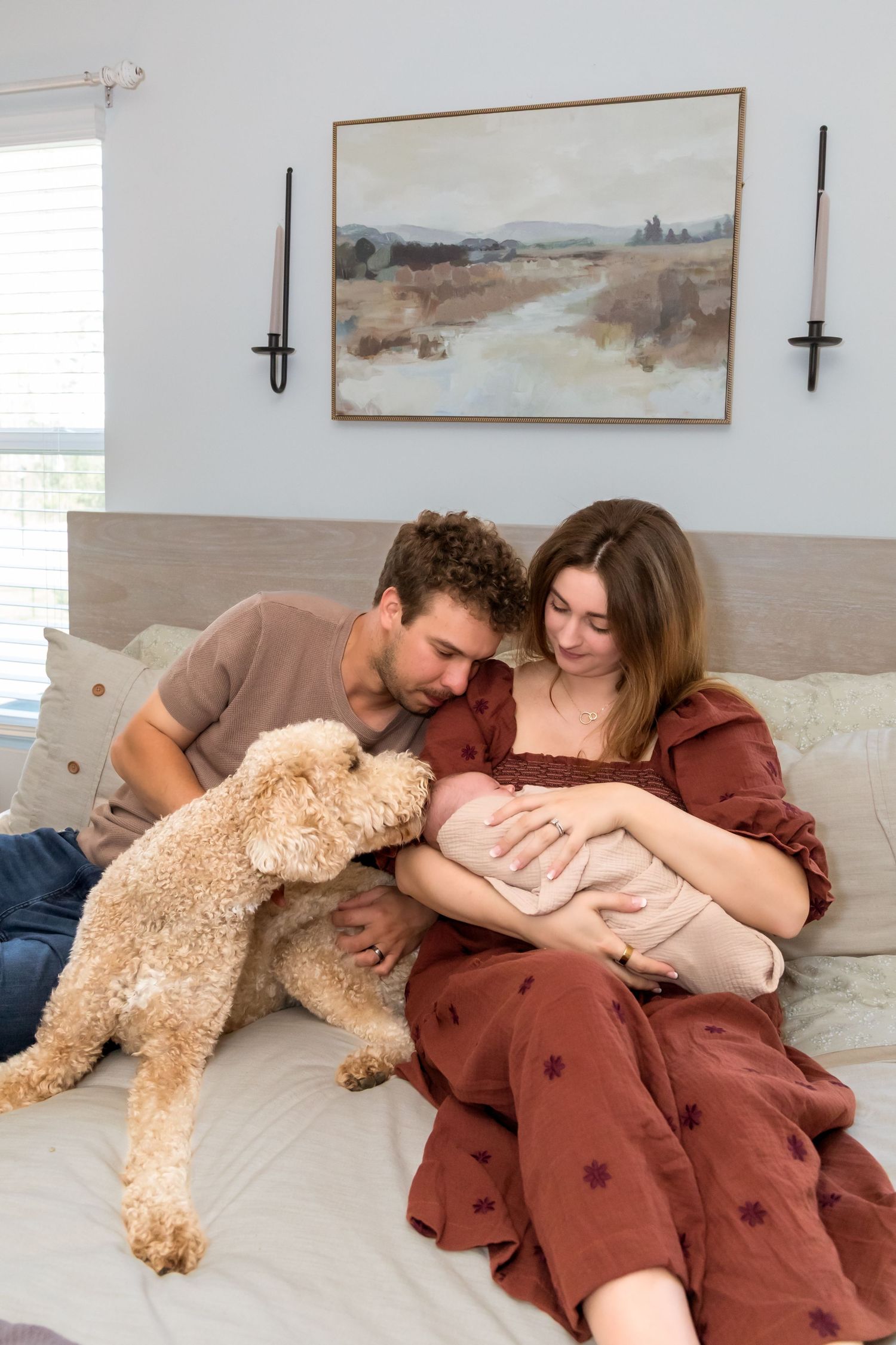New parents and their golden doodle dog cuddle with newborn baby on bed.
