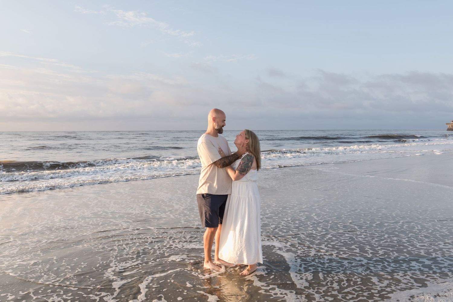 Couple embracing at the ocean's edge during golden hour, waves washing over their feet on a serene beach.