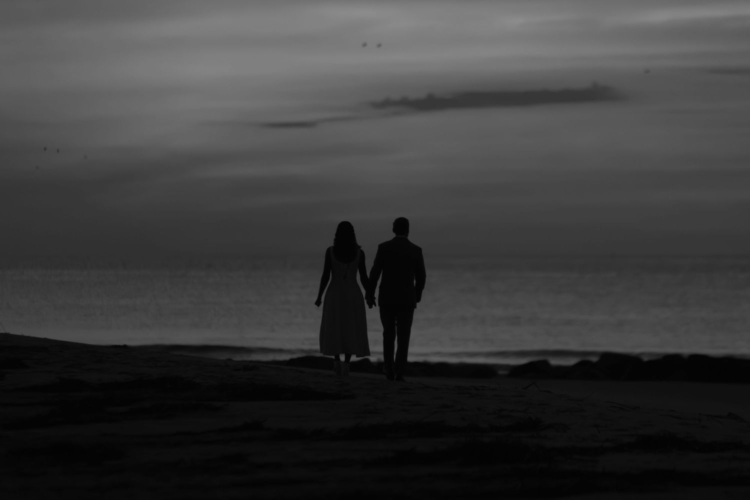 Silhouette of a couple holding hands on a beach at dusk in black and white photography.