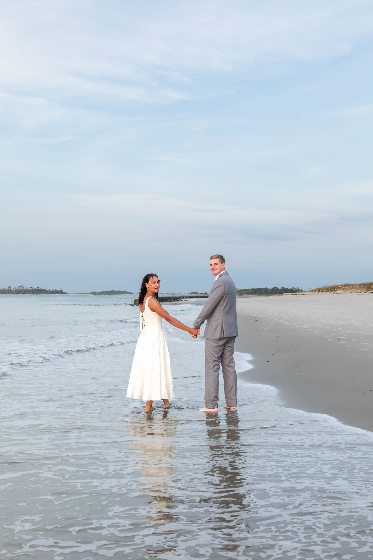 Bride and groom holding hands barefoot in ocean waves on a sandy beach in wedding attire.