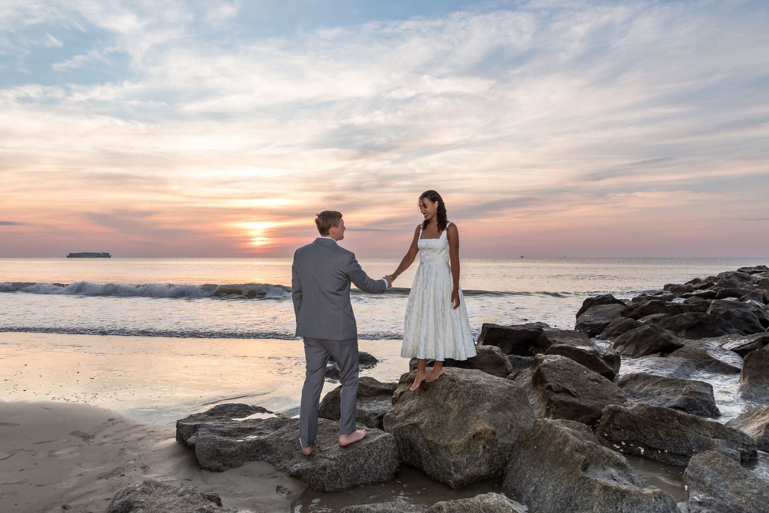 Couple holding hands on rocky beach at sunset, man in gray suit, woman in white dress, ocean backdrop.
