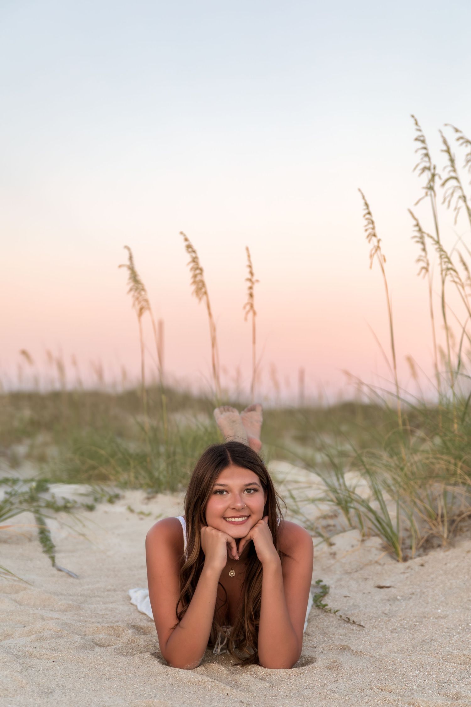 Young woman lying on sandy beach at sunset, smiling with hands under chin, sea oats and pink sky in background.