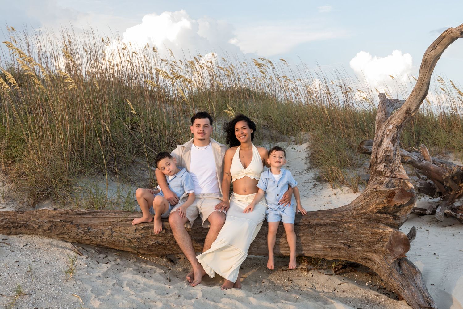 Family of four posing on driftwood at a sandy beach with sea oats in the background.