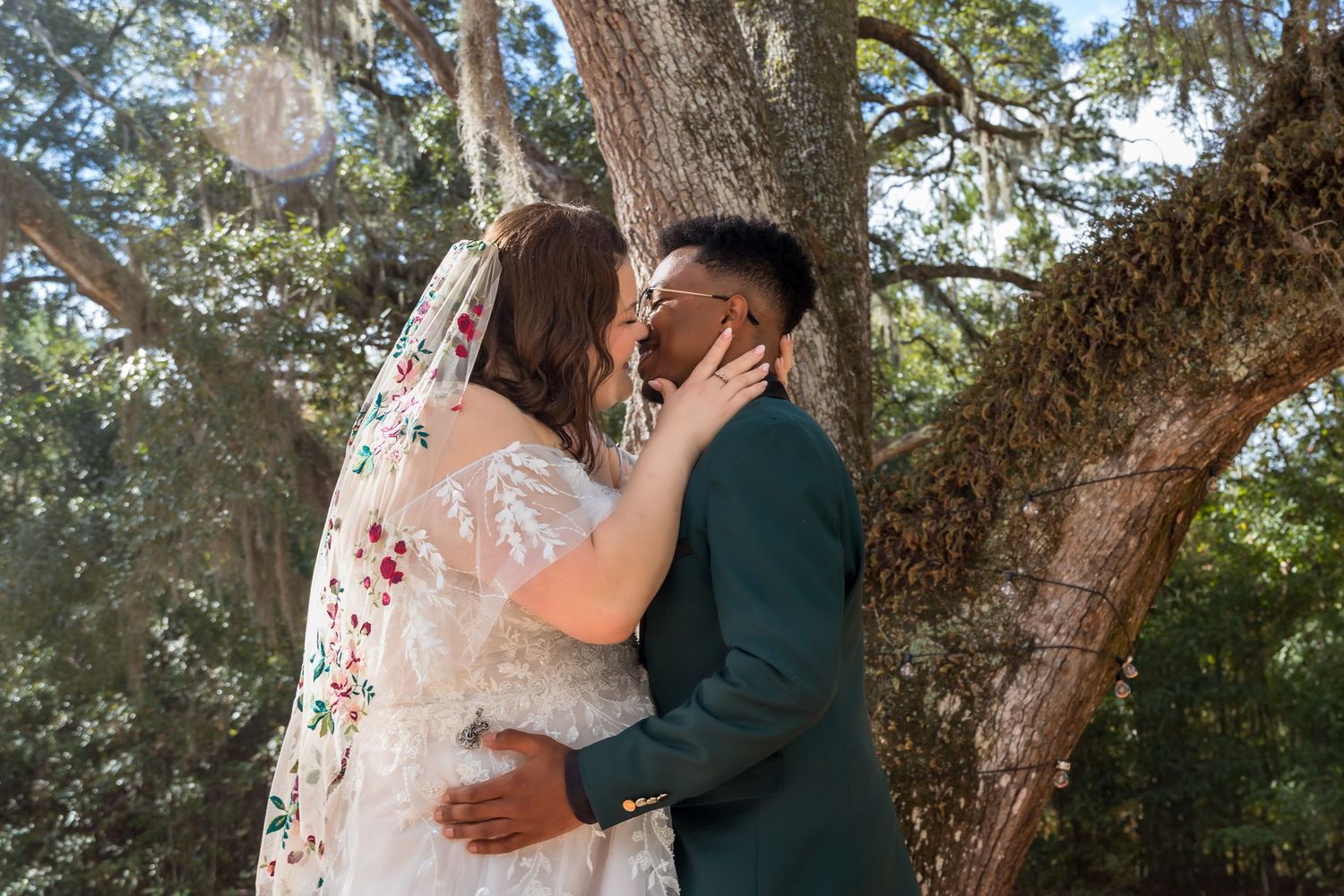 A couple shares a romantic kiss in a lush outdoor setting, bride in floral gown and groom in teal suit.