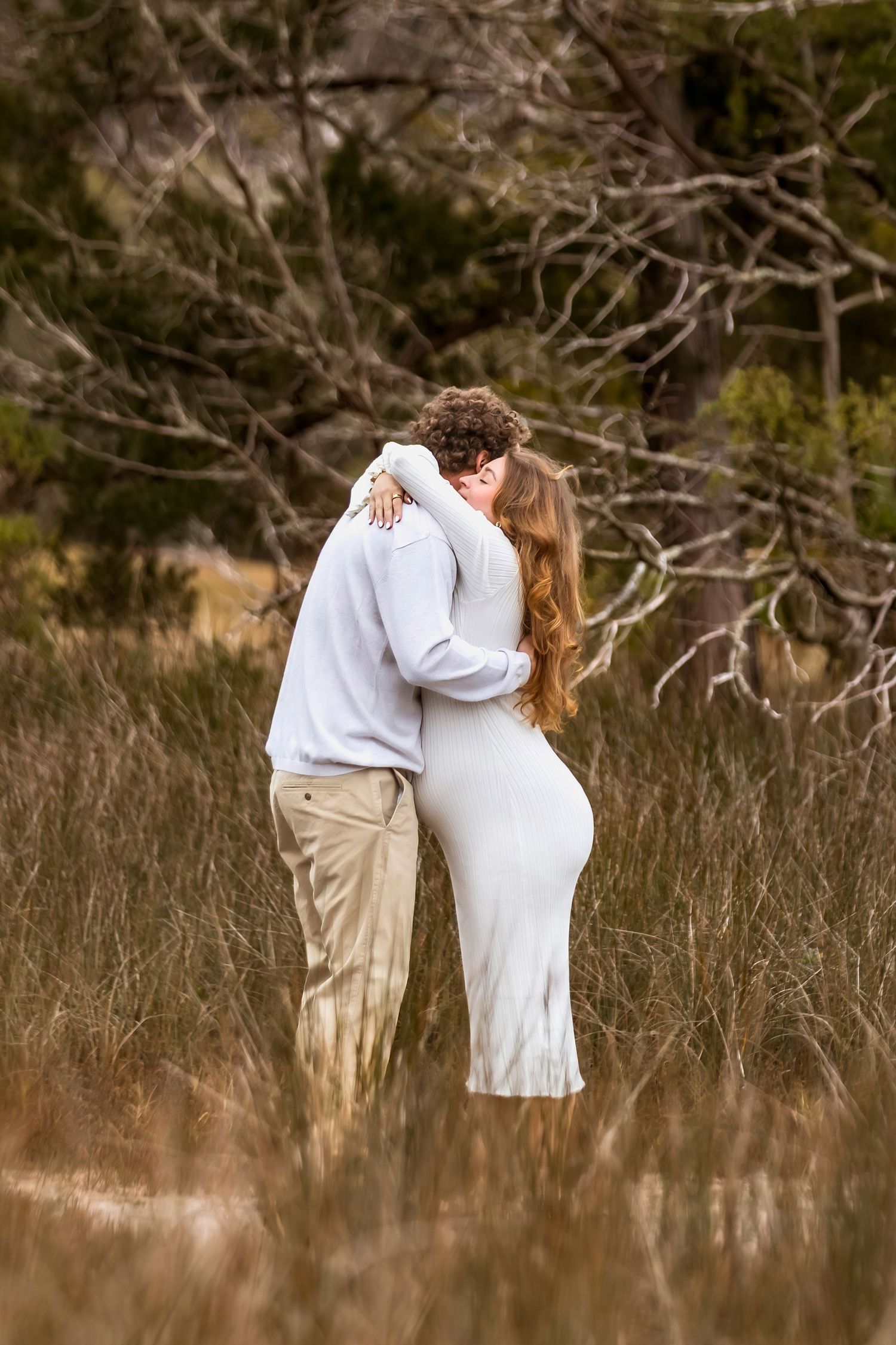 A romantic couple embraces in a field of tall grass during a sunset photoshoot.