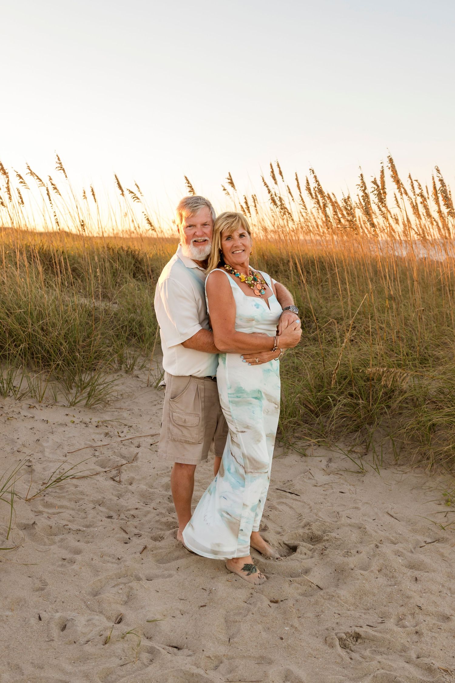 Couple embracing on a sandy beach with sea oats glowing in golden sunset light.