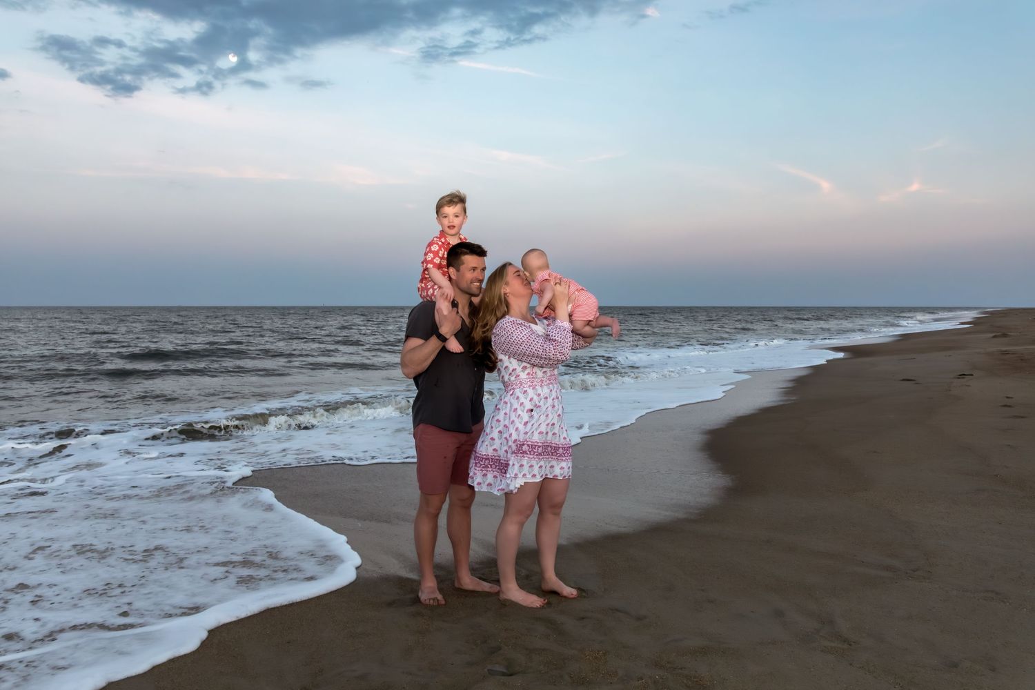 Family of four enjoying a sunset beach walk, with dad carrying son on shoulders and mom holding daughter in pink dress.
