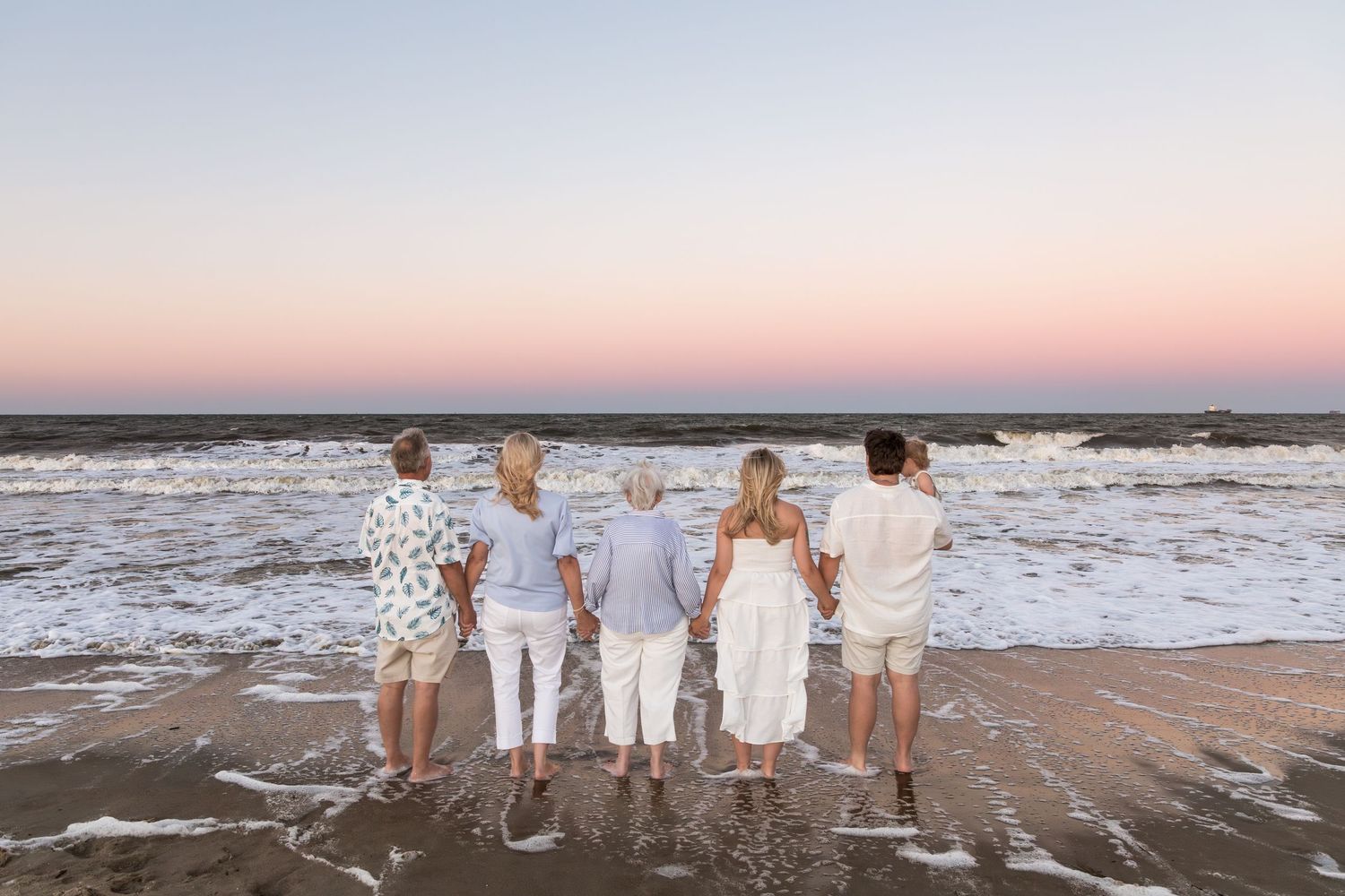 Five people holding hands facing ocean waves at sunset on a sandy beach during golden hour.