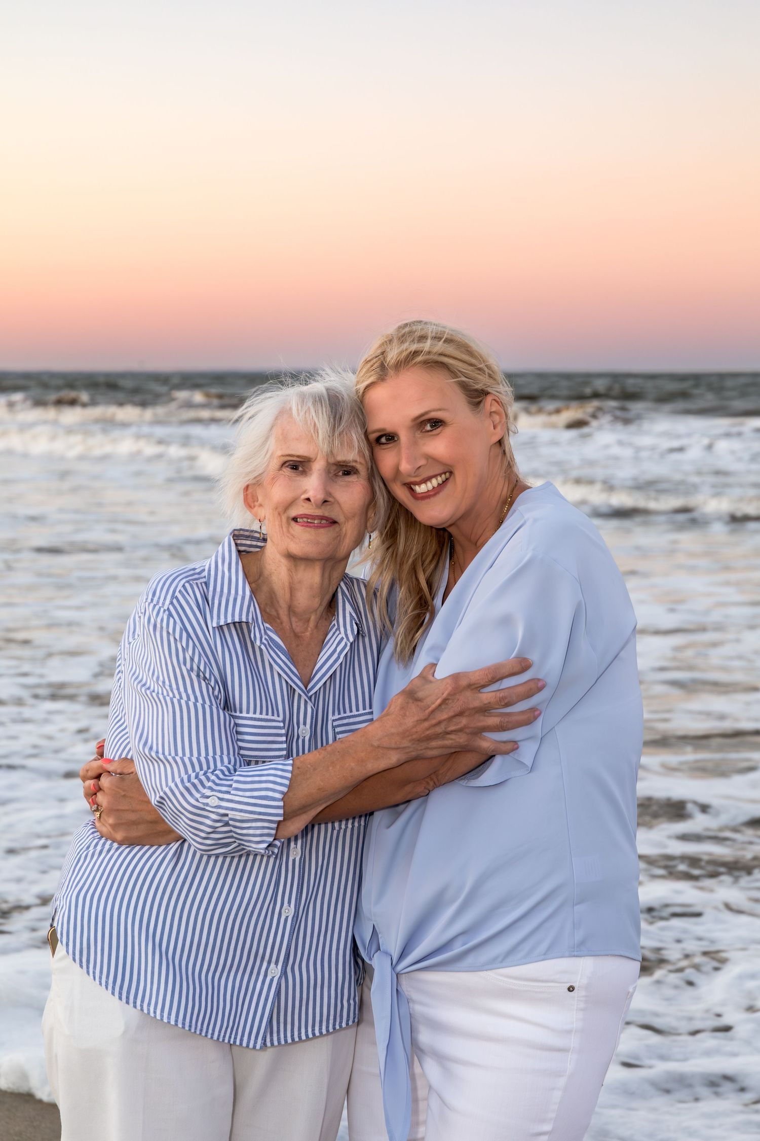 Two women embracing and smiling on beach at sunset with pink sky behind them.