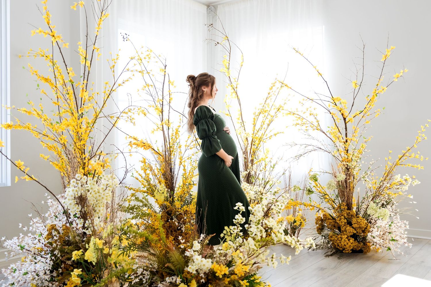 Woman in green dress surrounded by yellow and white floral arrangements in a bright white studio setting.