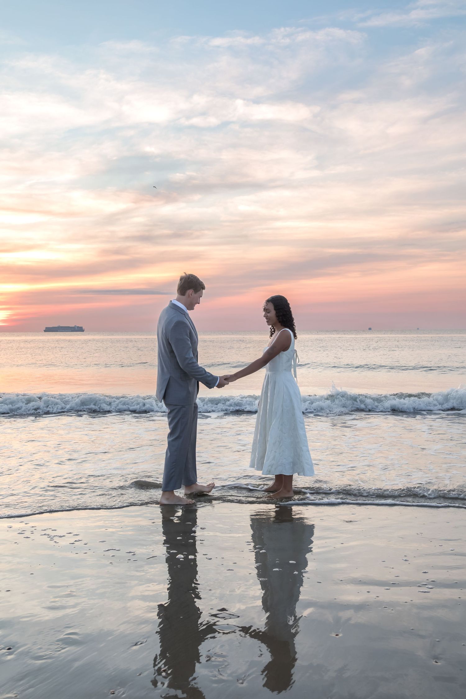 Couple holding hands on beach at sunset, wearing white dress and gray suit, standing in shallow ocean waves.