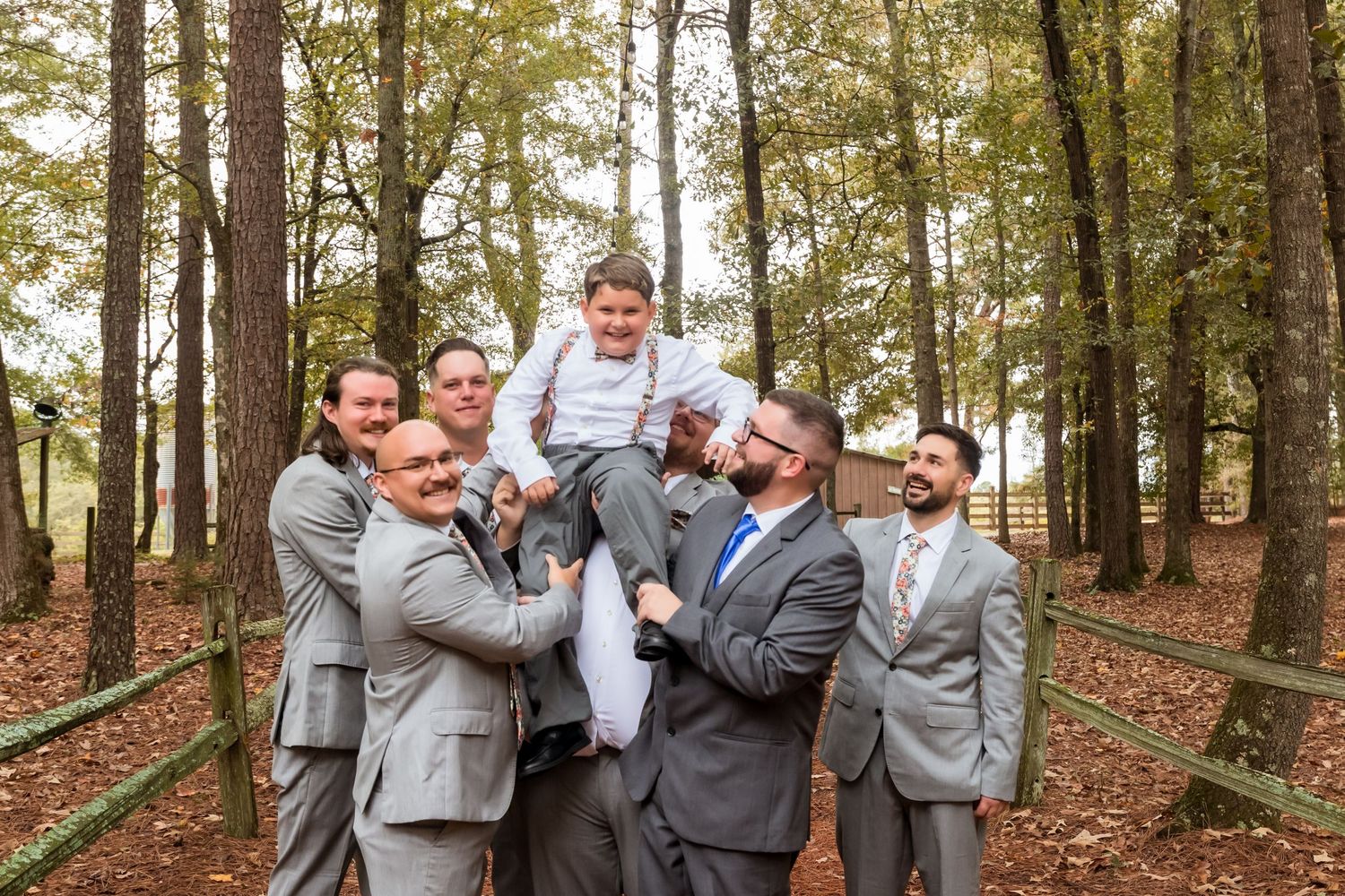Groomsmen in light gray suits share humorous moment in wooded outdoor setting.