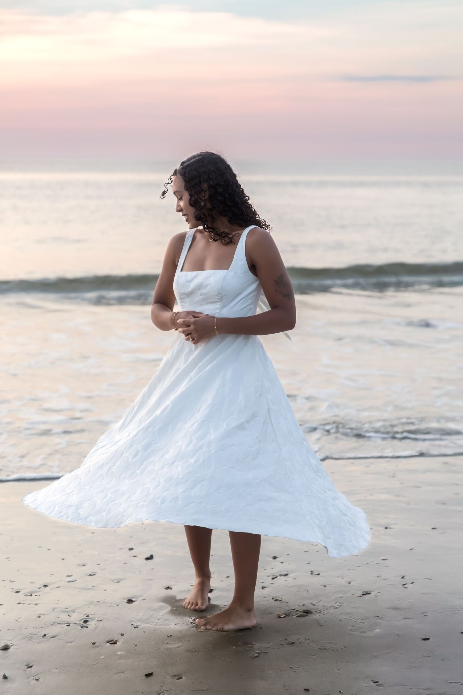 A barefoot woman in a flowing white dress stands on a sandy beach at sunset with ocean waves behind her.
