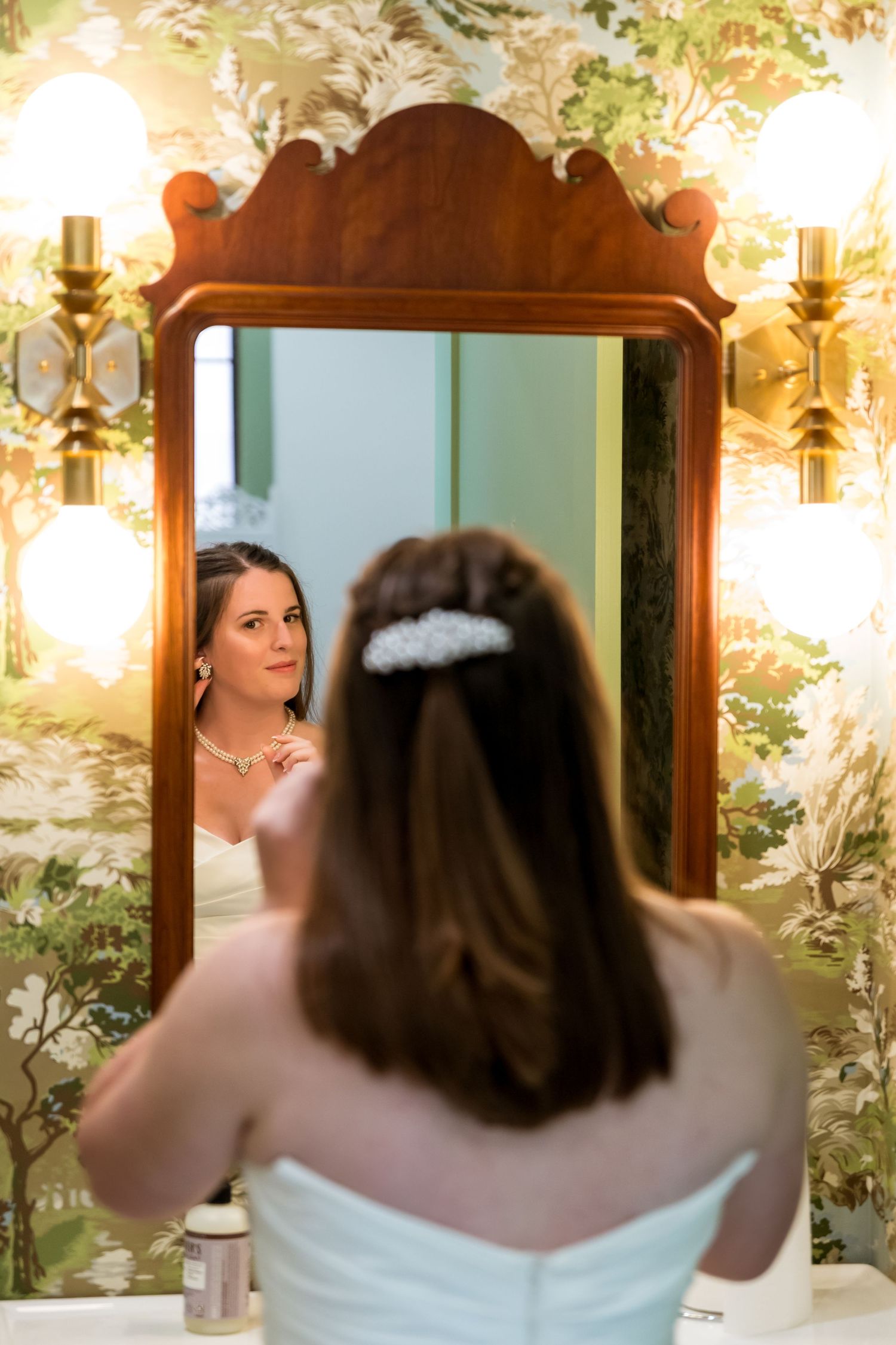 A reflection in an ornate wooden mirror shows a bride in a white dress with floral wallpaper and sconces in the background.