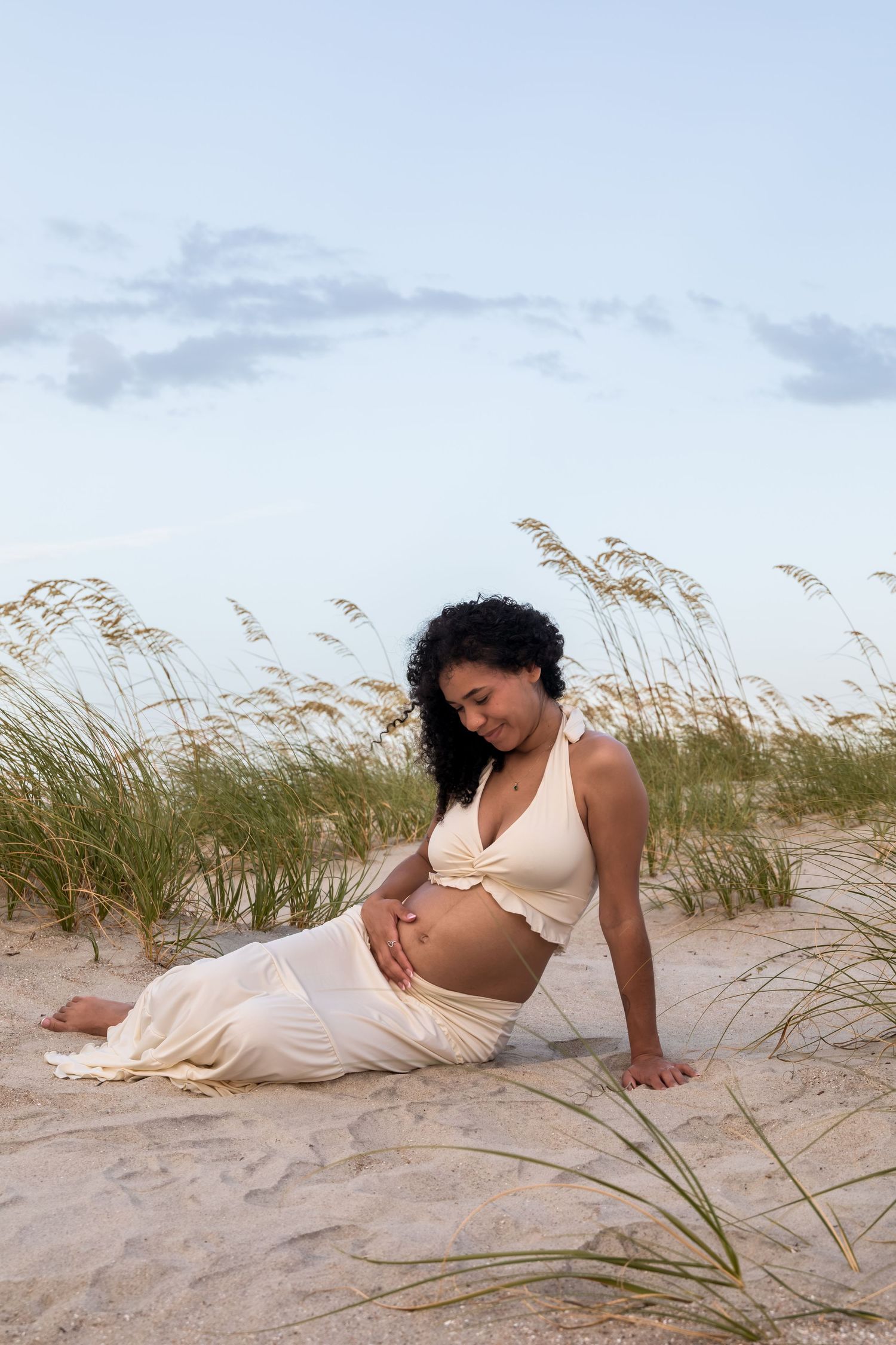 Pregnant woman in cream outfit sitting on beach sand dunes with sea oats, cradling her baby bump.