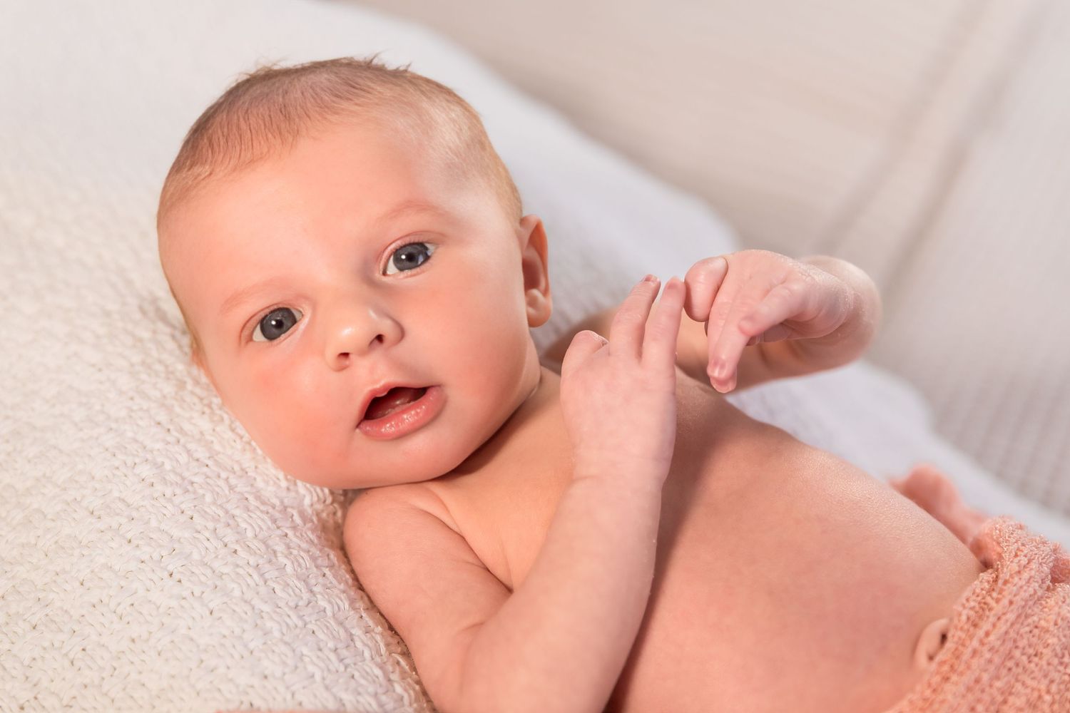 Newborn baby lying on a white blanket, holding an adult's finger with a curious expression.
