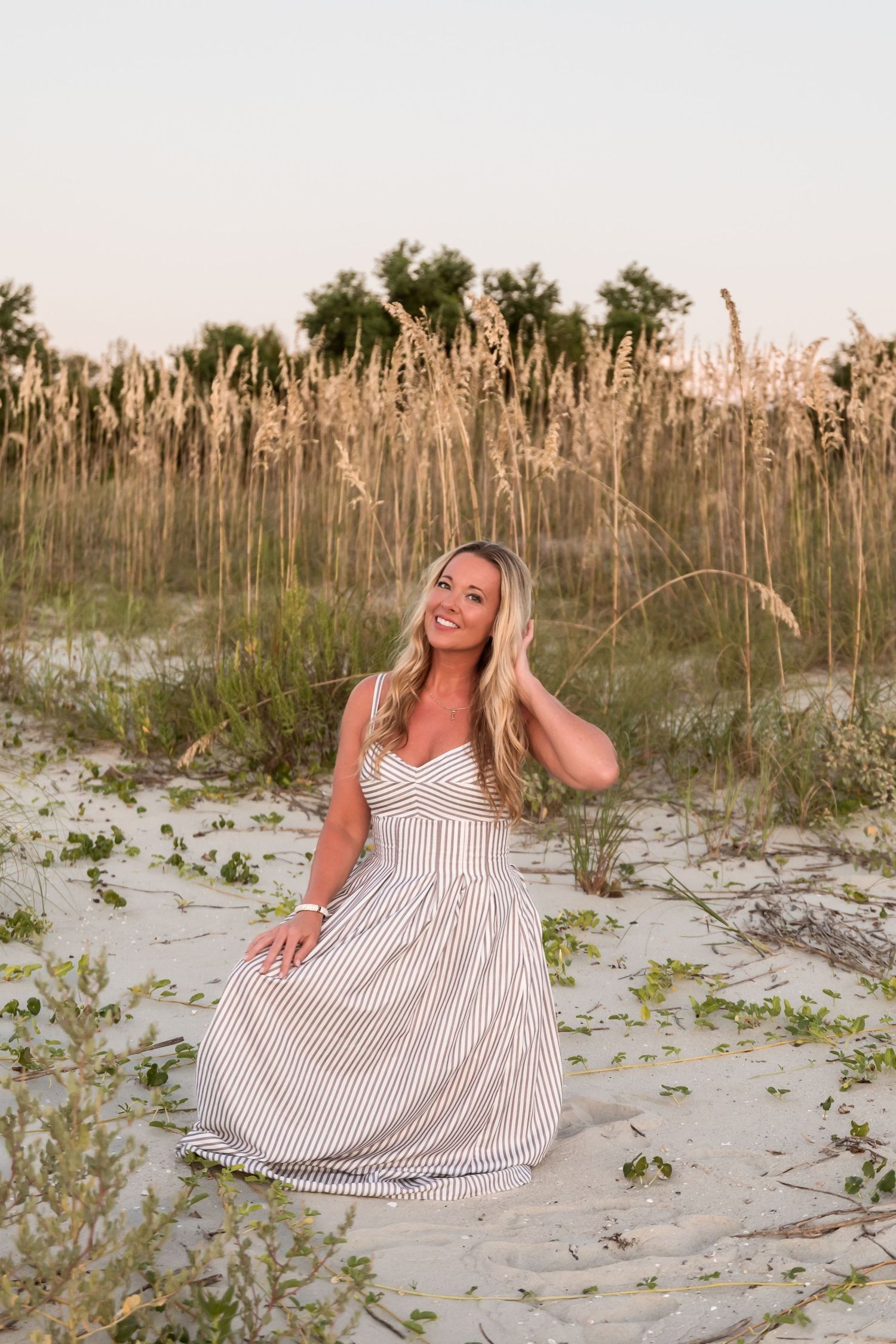 Smiling blonde woman in striped maxi dress sitting on sandy beach dunes with golden sea oats at sunset.