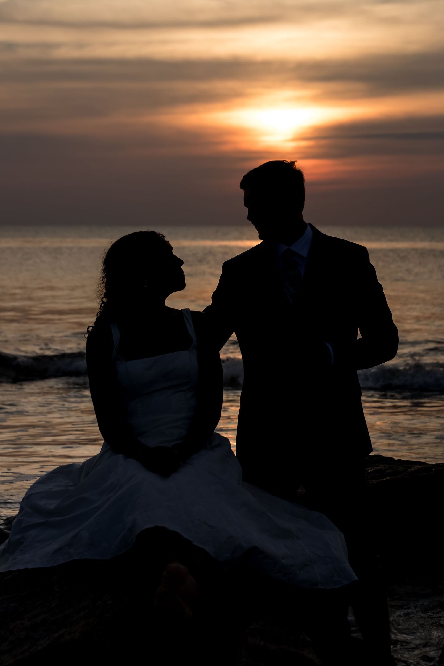 Silhouette of bride and groom sitting on rocks against dramatic ocean sunset.