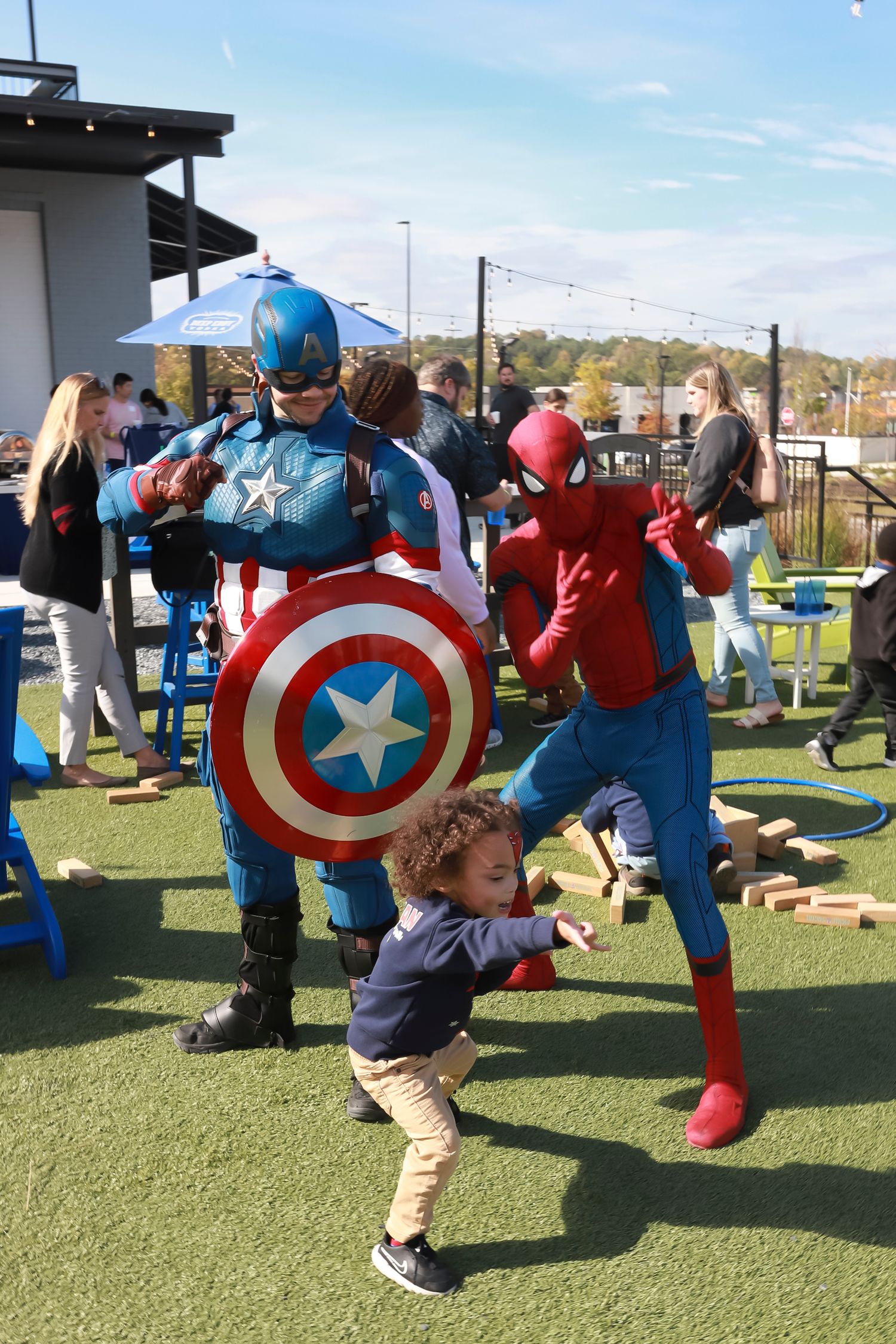 Children play with Captain America costumes and shields on a green outdoor playground surface.