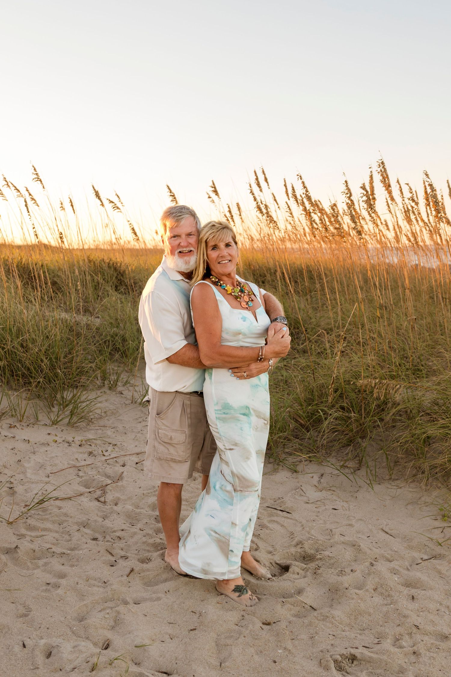 Couple embracing on sandy beach at sunset with sea oats in background.