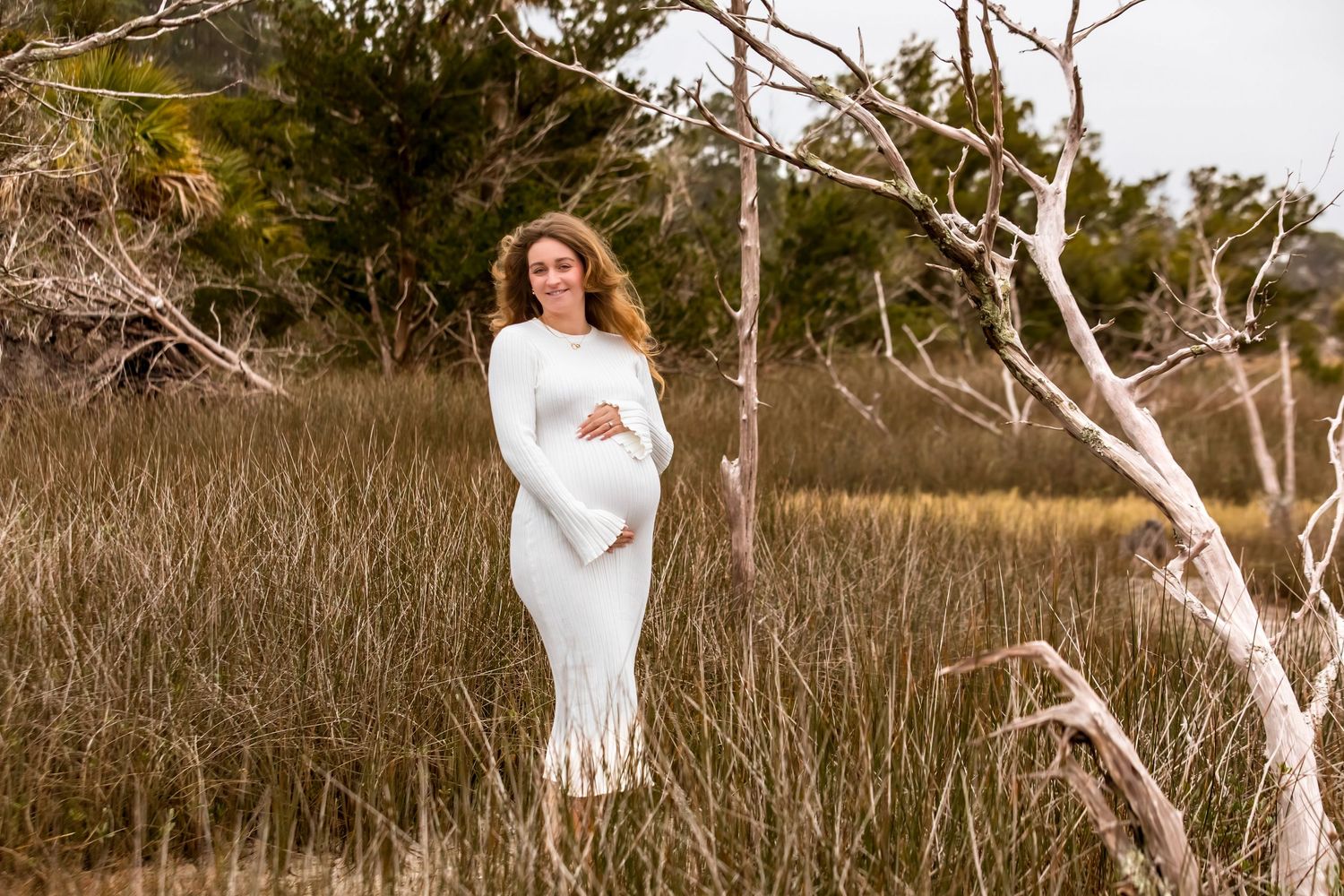 A woman in a white dress poses gracefully in a golden field during sunset with birch trees in the background.