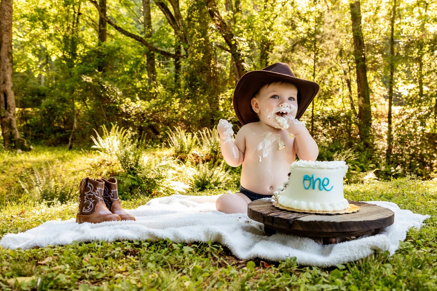 A child wearing a white dress and blue bow sits on a blanket in a sunlit forest for a portrait session.