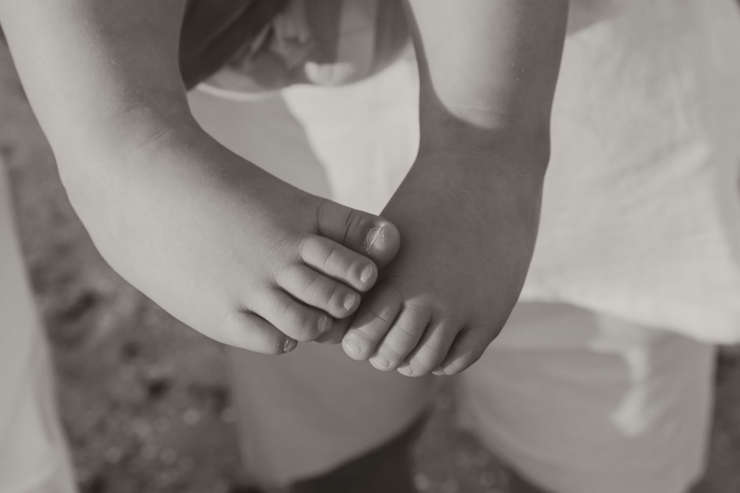 Black and white close-up of a child's bare feet held gently by an adult's hands.