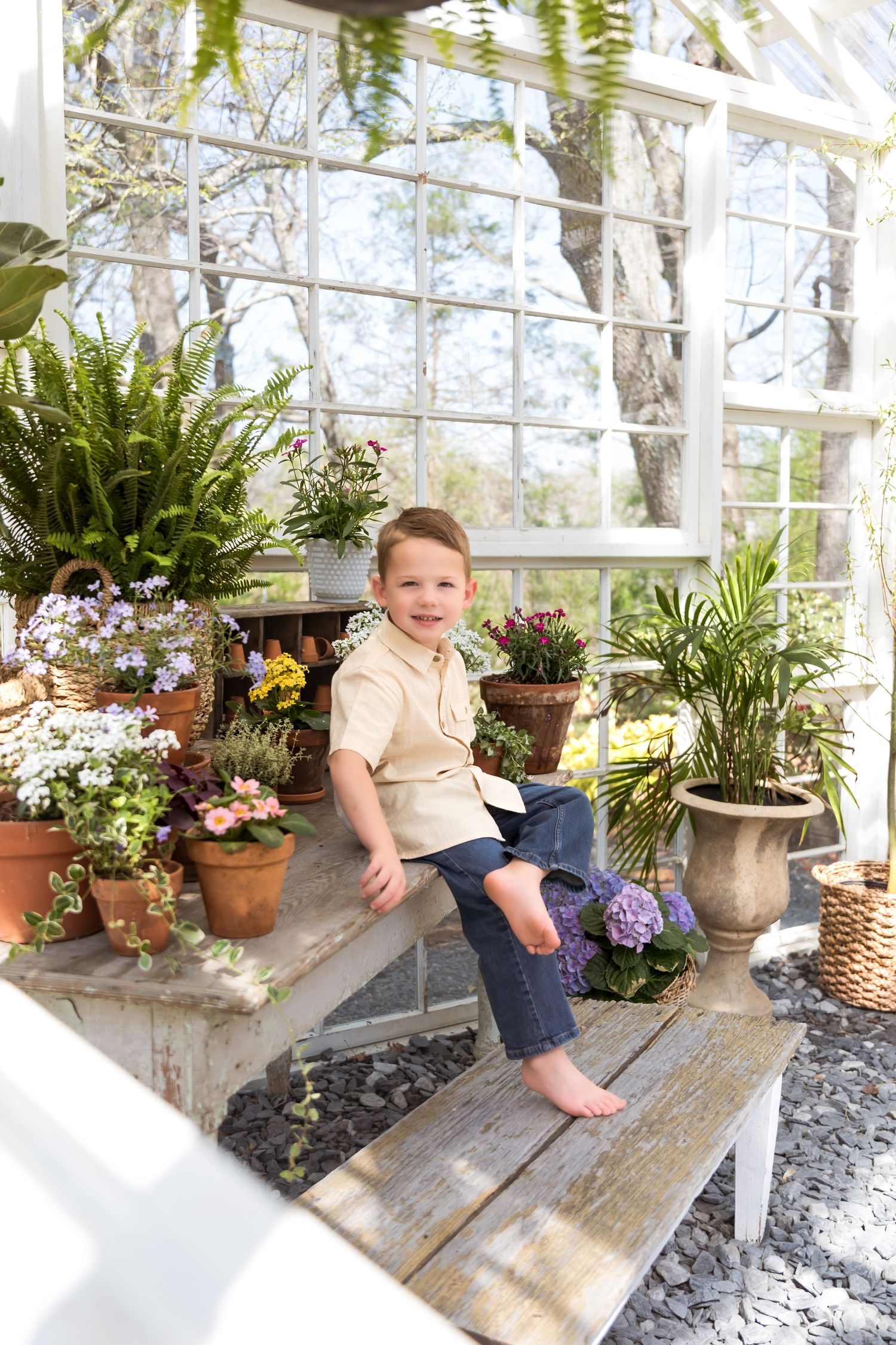 Young boy sitting barefoot on a wooden bench surrounded by colorful potted plants in a bright greenhouse.
