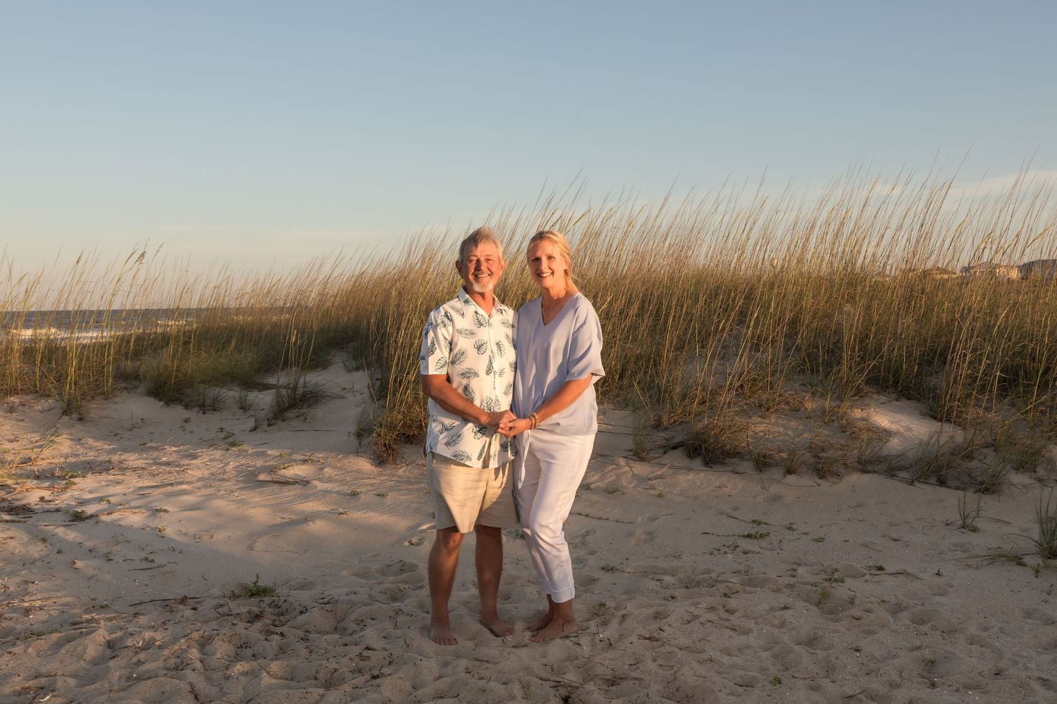Elderly couple smiling and holding hands on a sandy beach with sea oats and dunes at sunset.