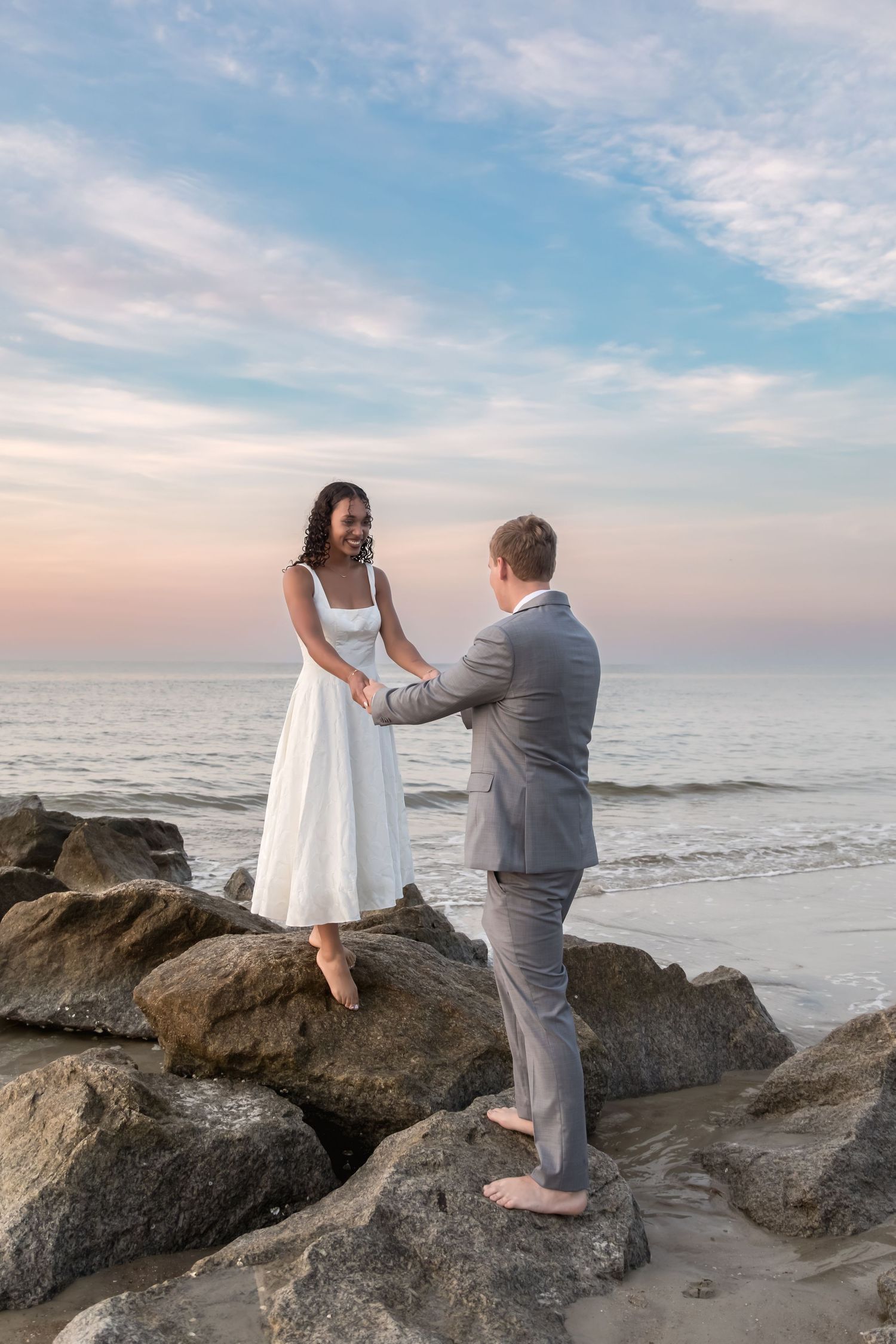 Couple in wedding attire holding hands on rocky beach at sunset, bride in white dress, groom in gray suit.