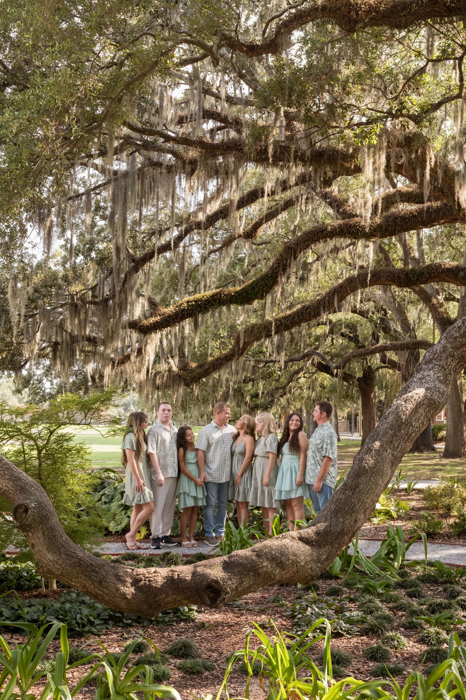 Family group portrait under a large moss-draped live oak tree in a lush Southern garden setting.
