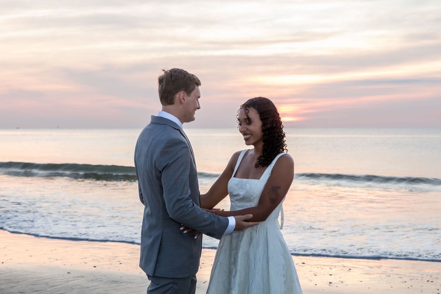 Couple holding hands on beach at sunset, man in gray suit and woman in white dress, romantic ocean backdrop.