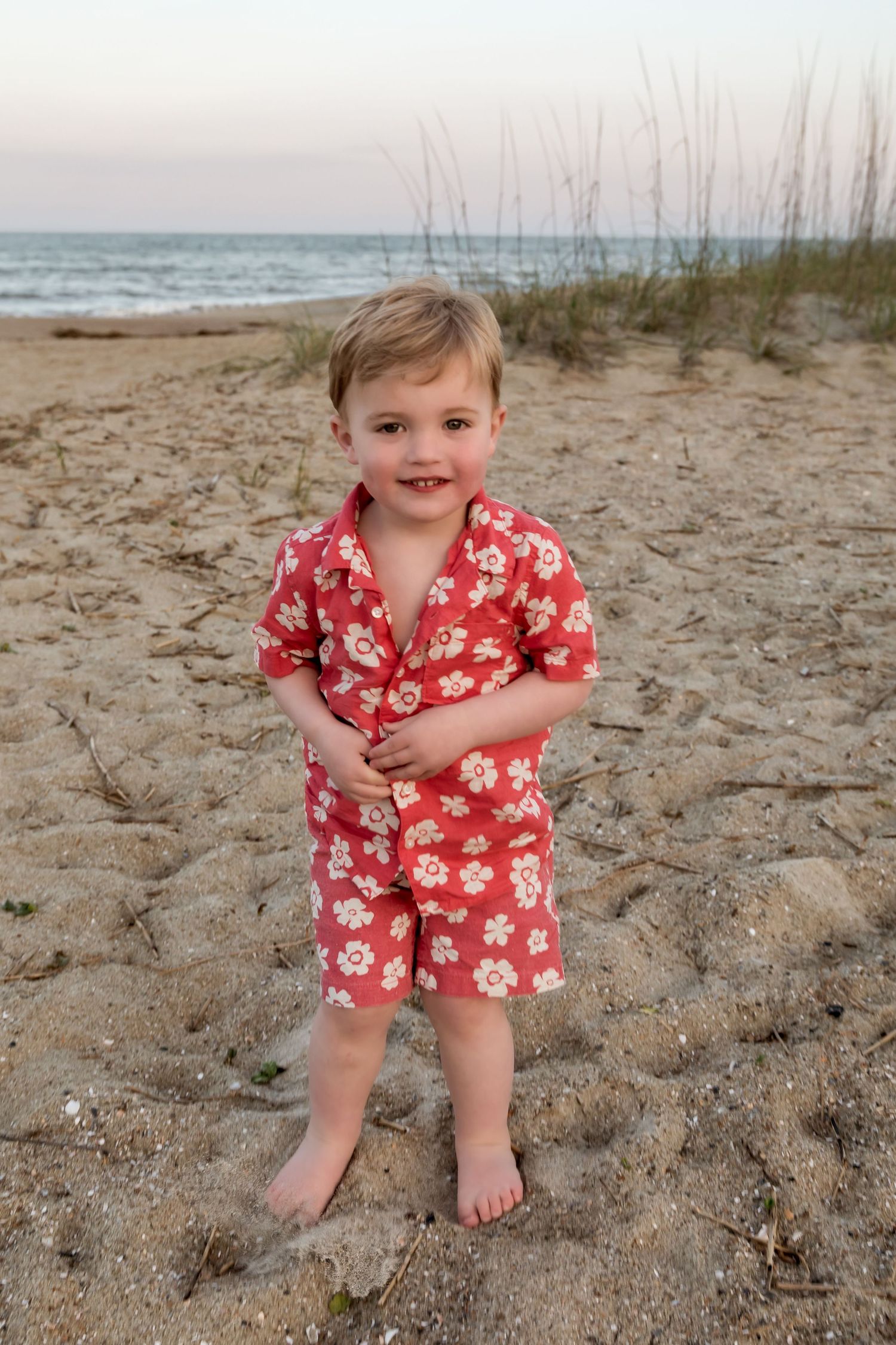 Adorable toddler boy in red floral matching shirt and shorts set smiling on sandy beach at dusk.
