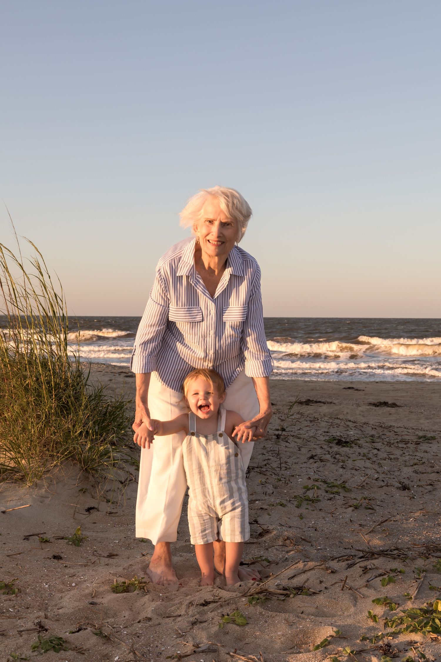 Grandmother holding toddler's hands on sandy beach with ocean waves in background.