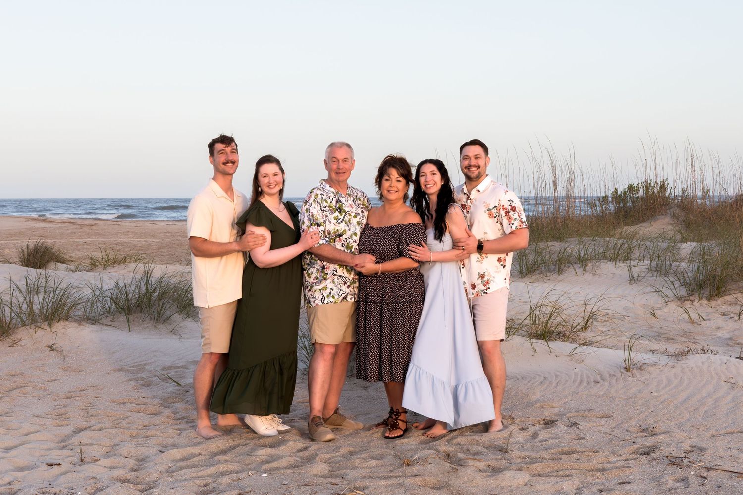 Family of six smiling together on a sandy beach at sunset, with ocean waves and dunes in the background.
