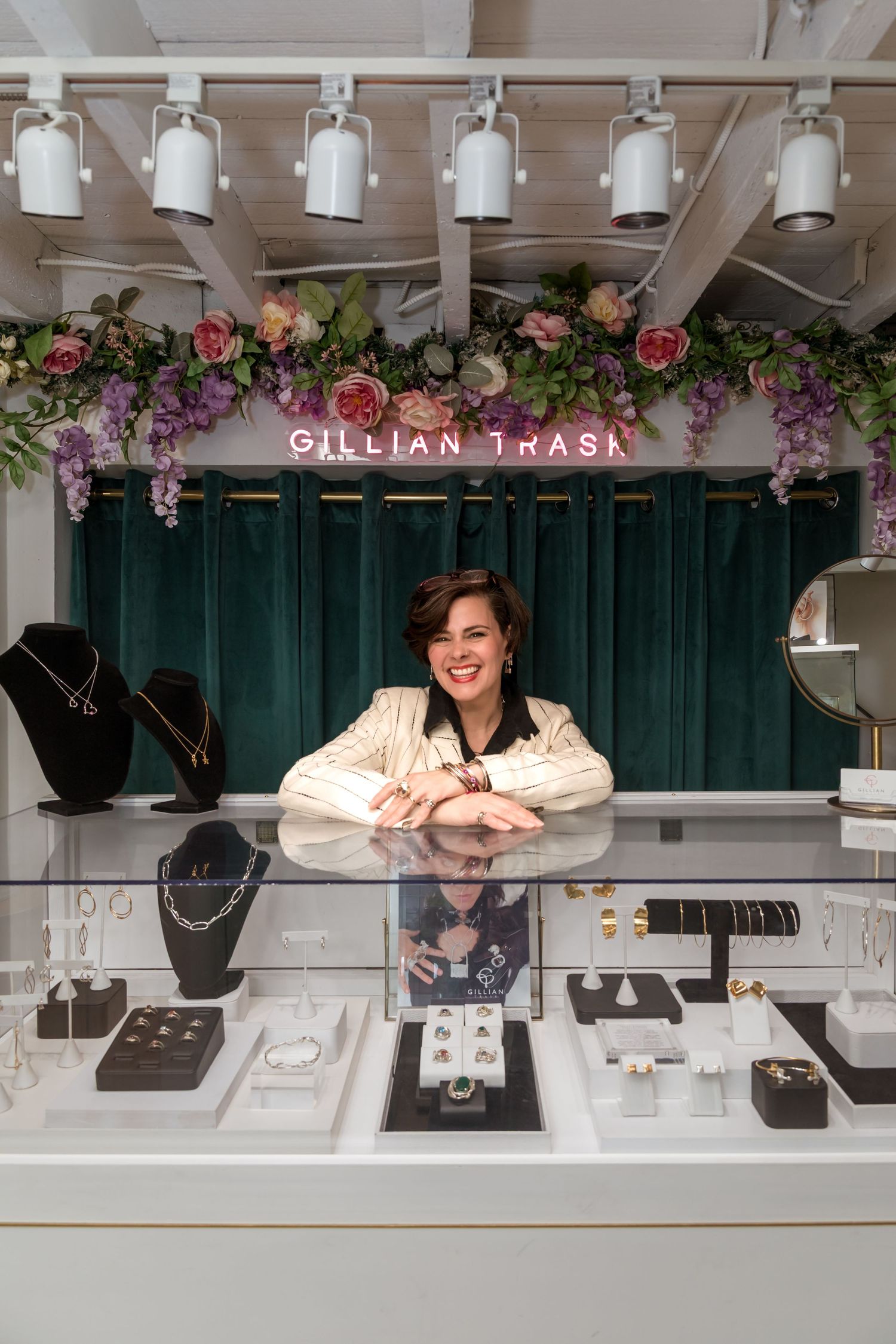 Woman smiling behind a jewelry display case at Gillian Trask boutique, surrounded by necklaces, rings, and earrings.