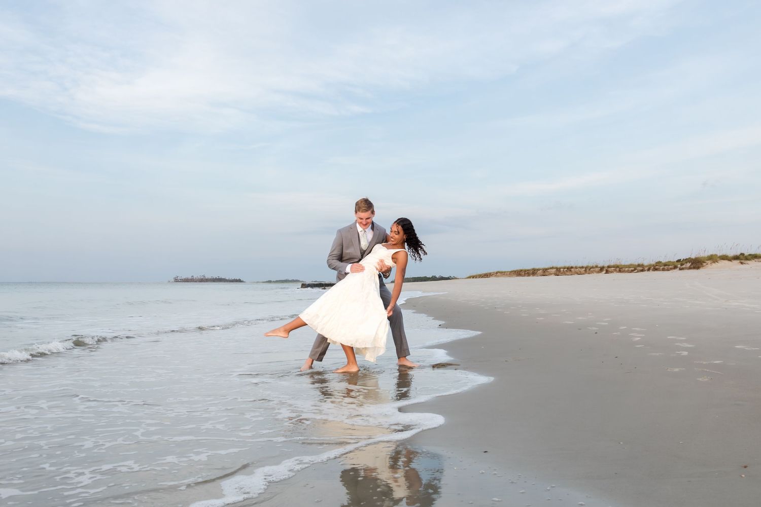Bride and groom dancing joyfully at the ocean shoreline during a romantic beach wedding celebration.