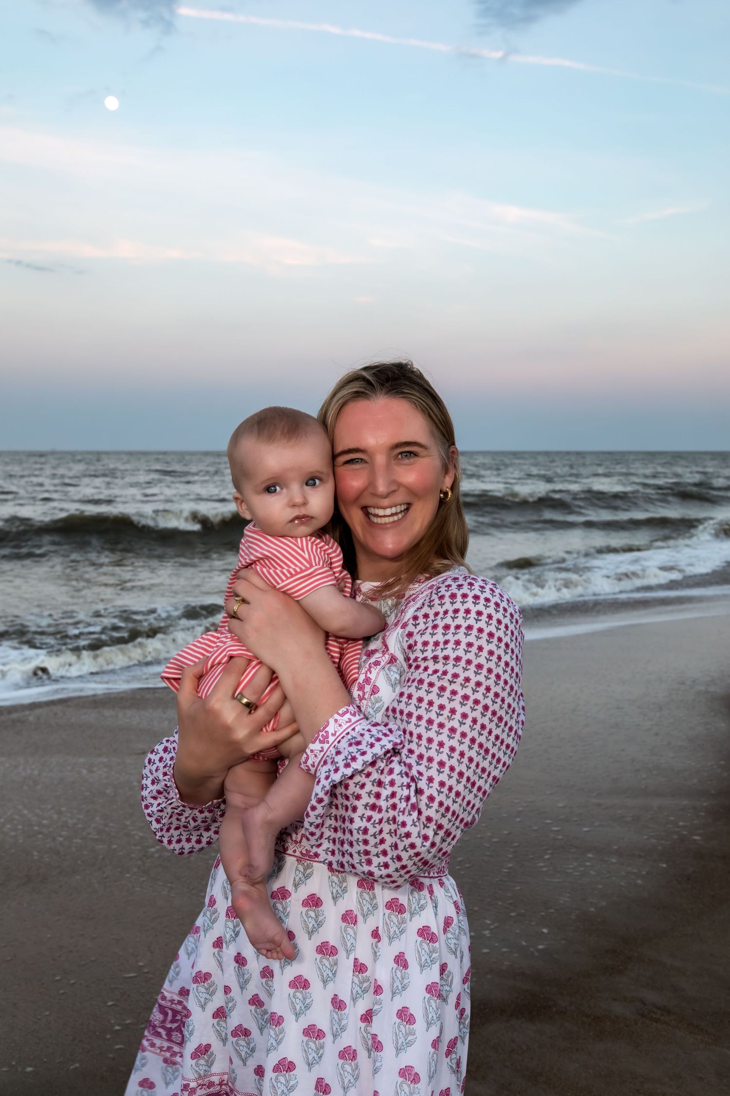 Smiling woman holding baby on beach at sunset, both wearing matching pink floral outfits with ocean waves behind them.