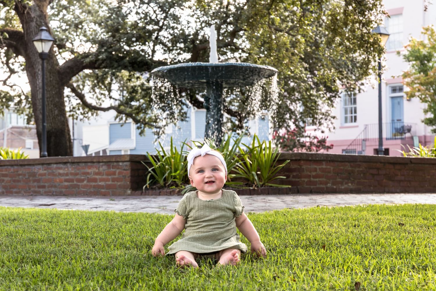 Happy baby sitting on green grass in front of a decorative fountain in a sunny park setting.