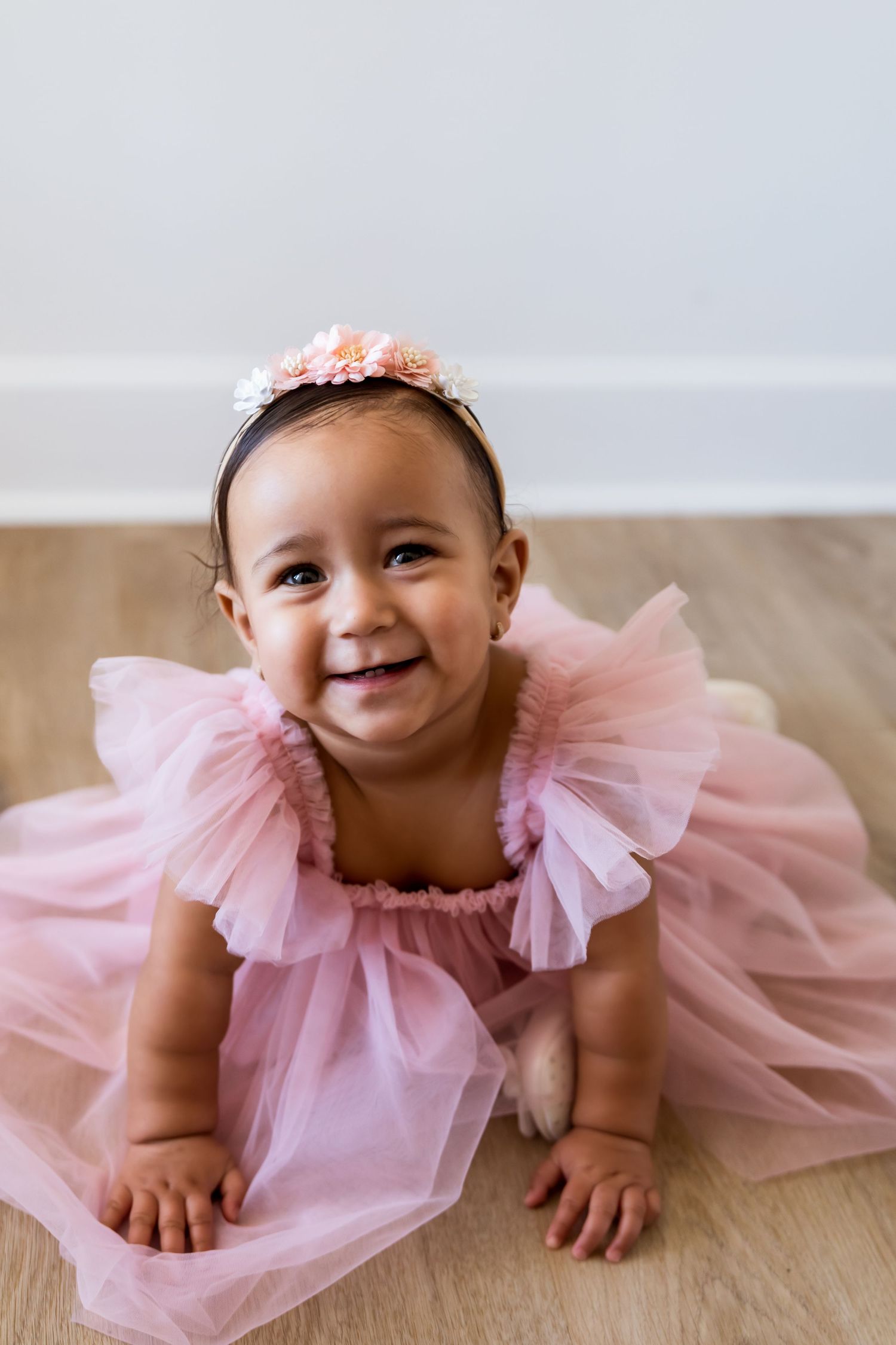 Baby wearing pink tulle dress and floral headband lies on wooden floor smiling in multiple poses.