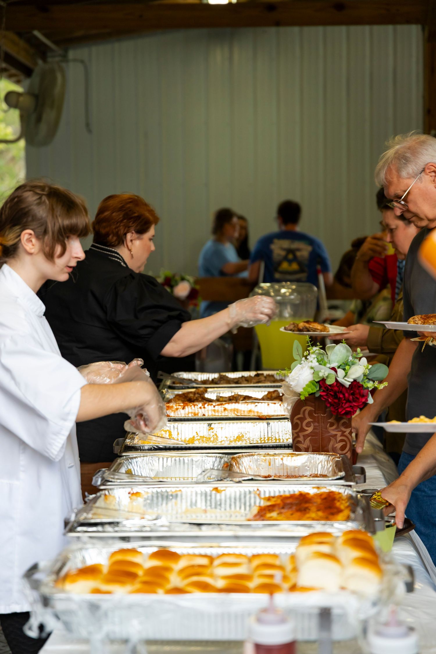 Long buffet table with catering trays and people serving food at a community event.
