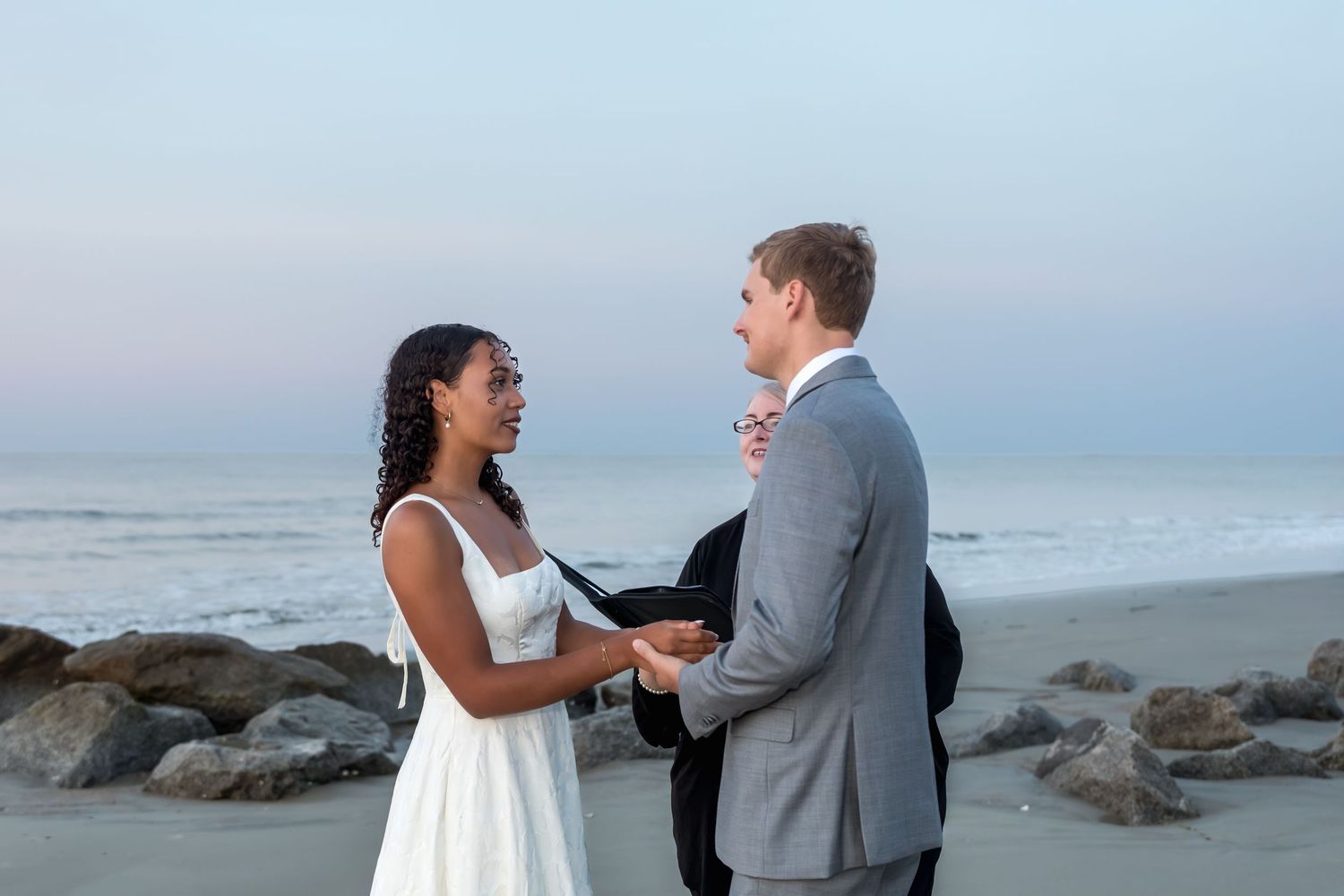 Couple exchanging vows on a beach at dusk, bride in white dress, groom in gray suit, officiant behind them.