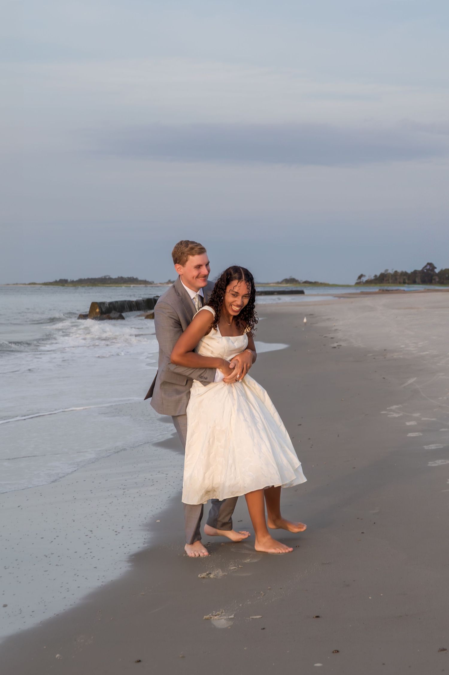 Groom in gray suit embraces laughing bride in white dress on beach at twilight.
